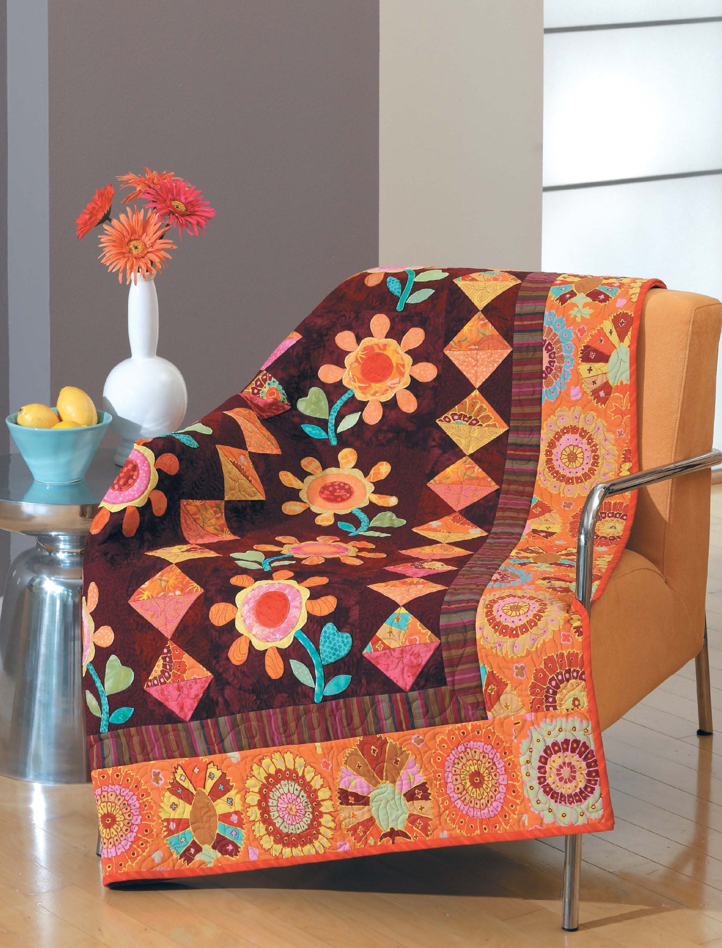 A vibrant floral quilt from Leisure Arts' "City Walks—10 Contemporary Quilt Designs" drapes over an orange chair beside a metal table with a vase of red flowers and a bowl of lemons.