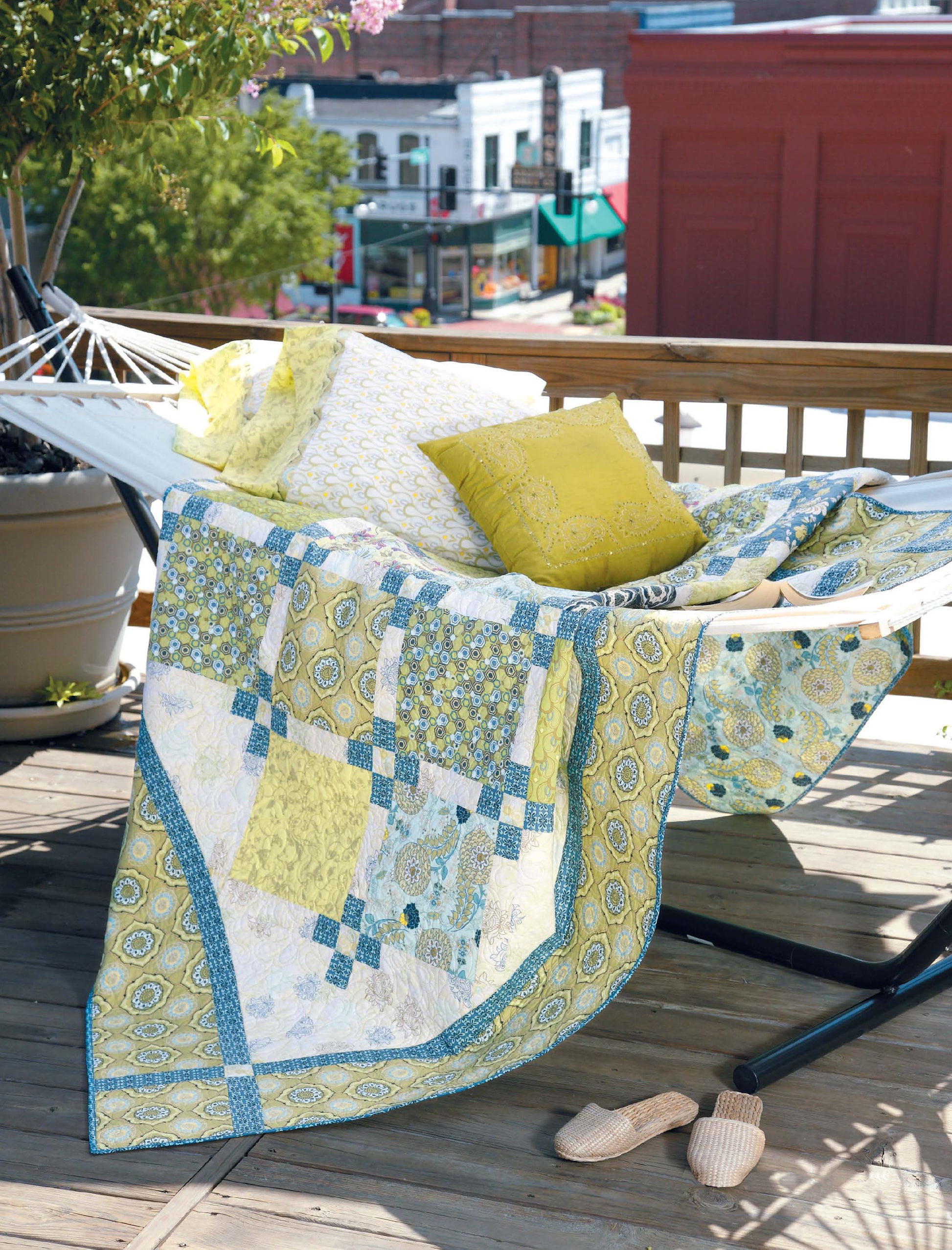 A hammock on a wooden deck holds a patterned quilt from Leisure Arts' City Walks—10 Contemporary Quilt Designs, plus a yellow pillow and folded blanket. Slippers rest nearby while trees and buildings line a small town street in the background.