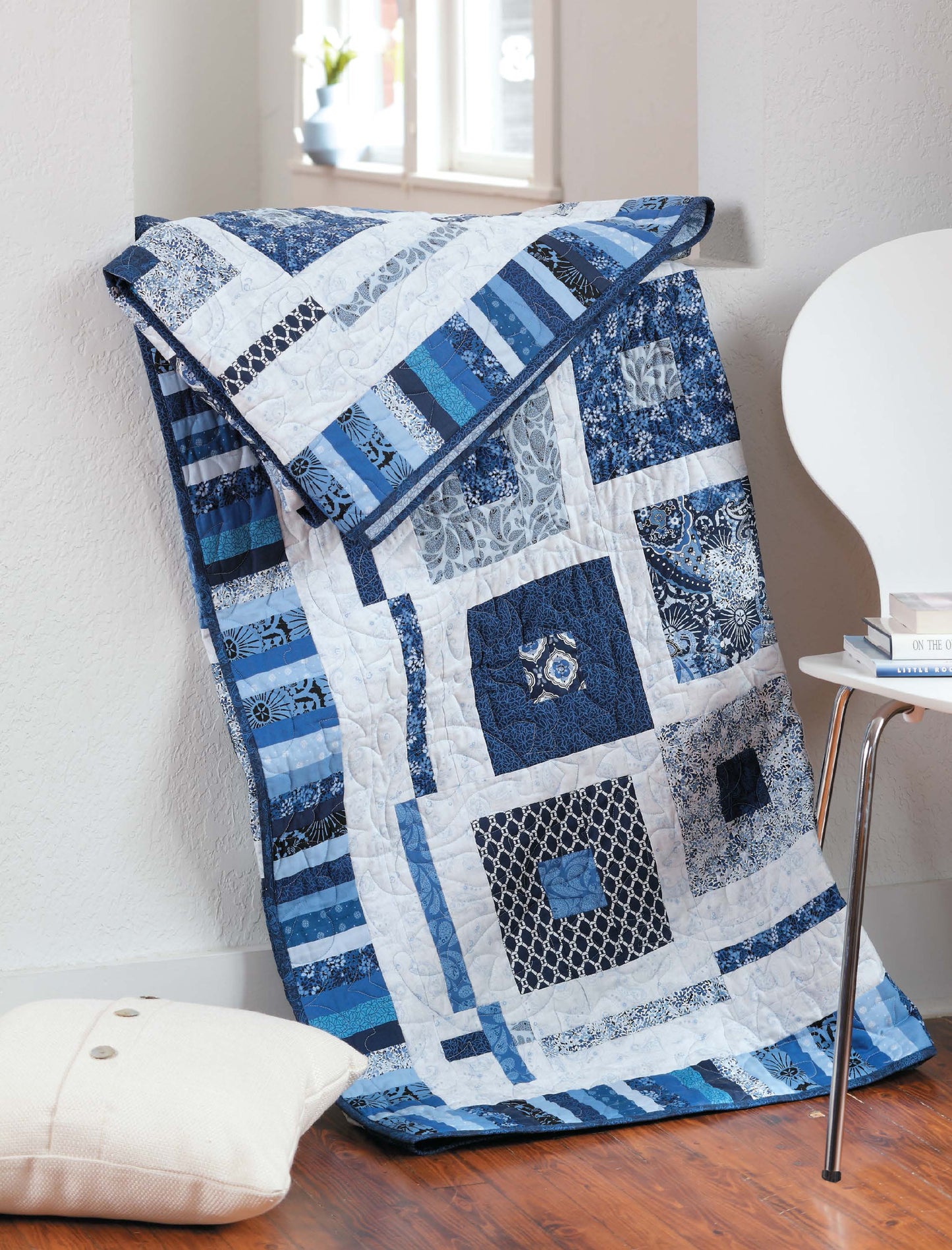 The City Walks—10 Contemporary Quilt Designs quilt by Leisure Arts, featuring blue and white geometric patterns, is draped over a white chair near a window on a wooden floor beside a white cushion.