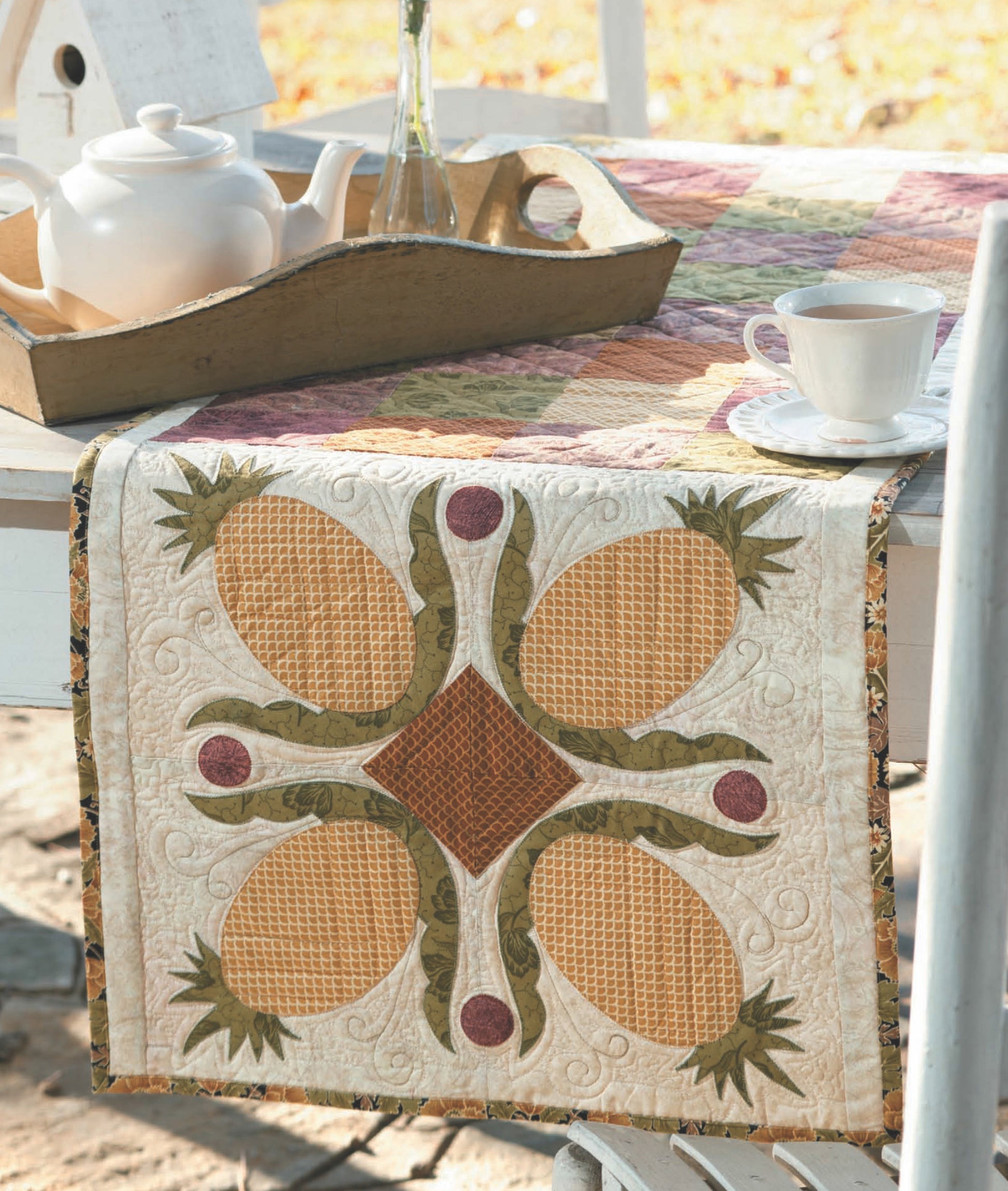 A white outdoor table displays the Leisure Arts Quilts to Warm Today's Home Digital Download, featuring a pineapple-themed quilted runner with applique techniques, plus a white teapot, glass vase, and teacup amid sunlight and autumn leaves.