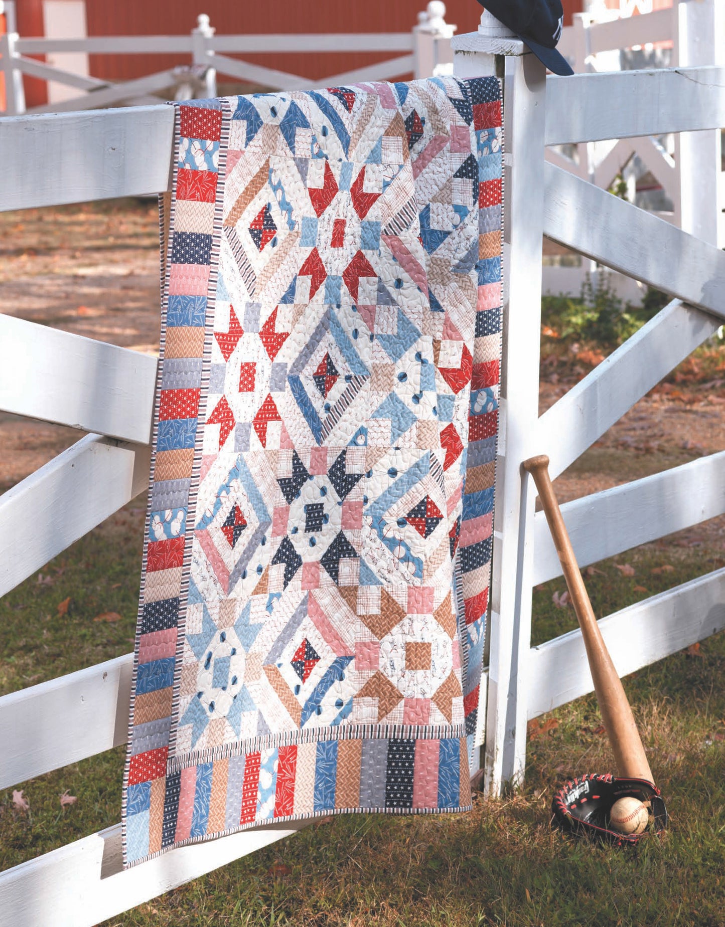 The "Quilts to Warm Today's Home Digital Download" by Leisure Arts features vibrant patchwork quilts with geometric patterns and applique techniques, perfect for draping over a white wooden fence as shown in the scene.