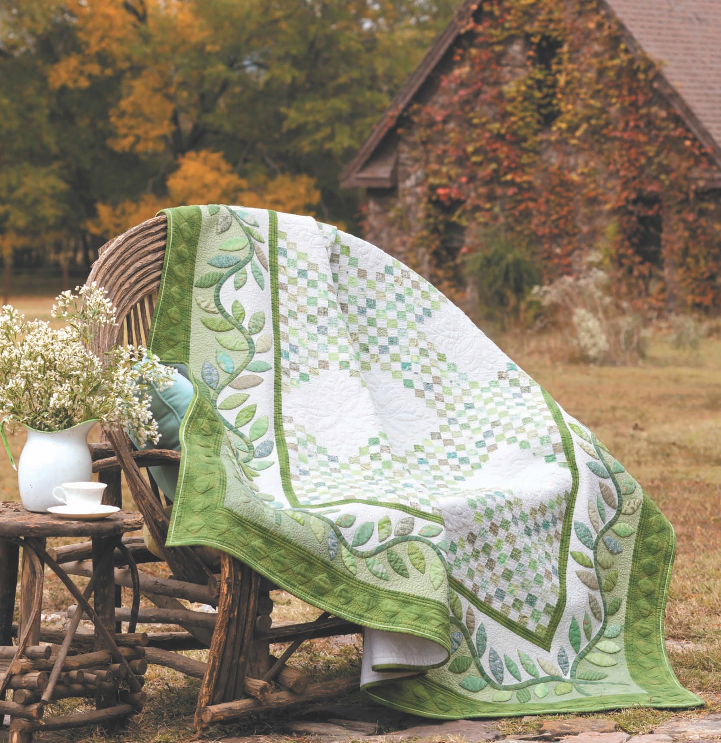 The Quilts to Warm Today's Home Digital Download by Leisure Arts features a green and white leaf-patterned homestyle quilt draped over a wicker chair beside a rustic wooden table, outdoors near an ivy-covered stone building.