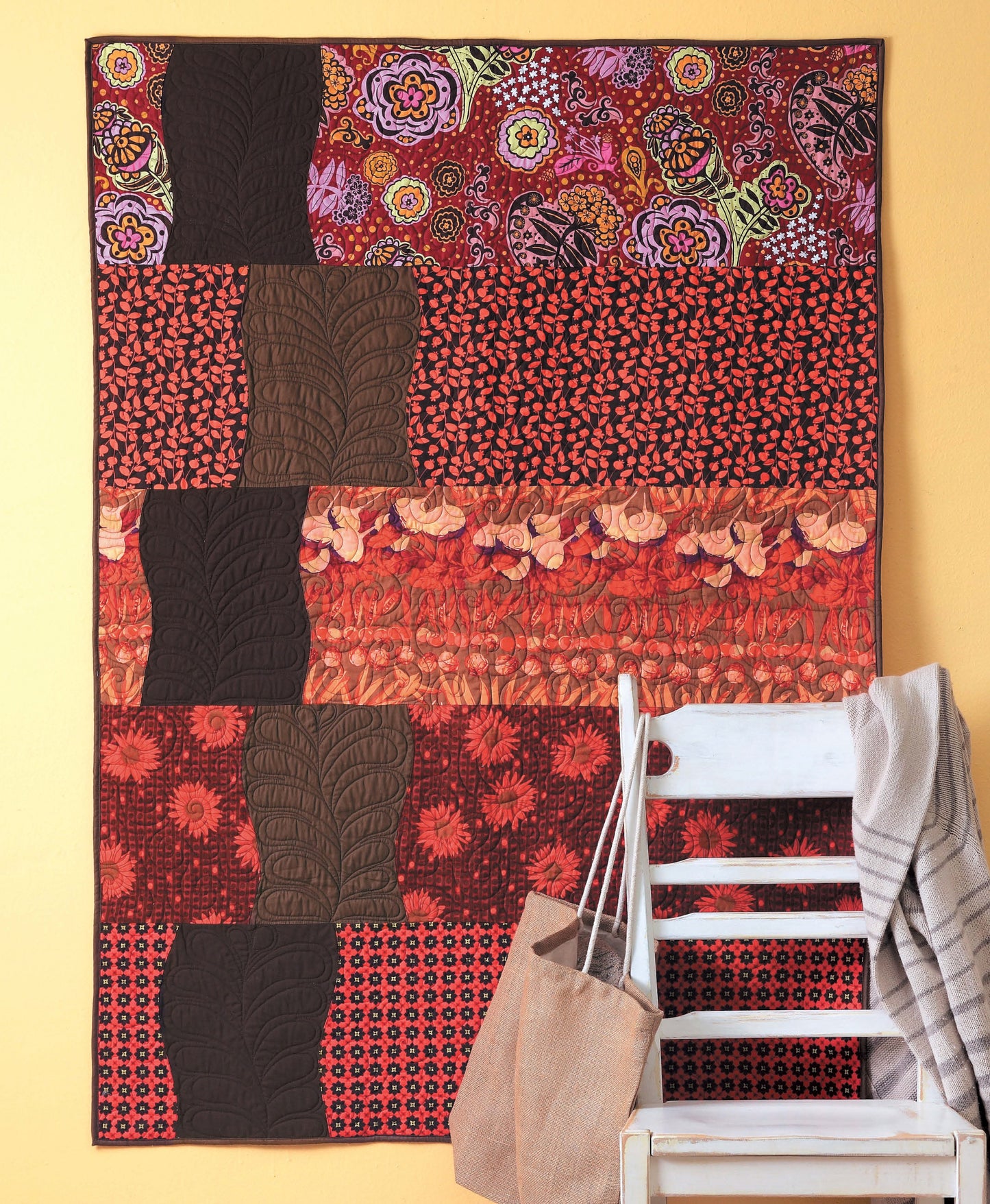A vibrant quilt made with the “Learn to Sew Easy Curves” pattern by Leisure Arts—featuring red, orange, and brown floral designs with gracefully curved seams—hangs on a yellow wall. In front of the quilt sits a white wooden chair holding a burlap bag and gray fabric.