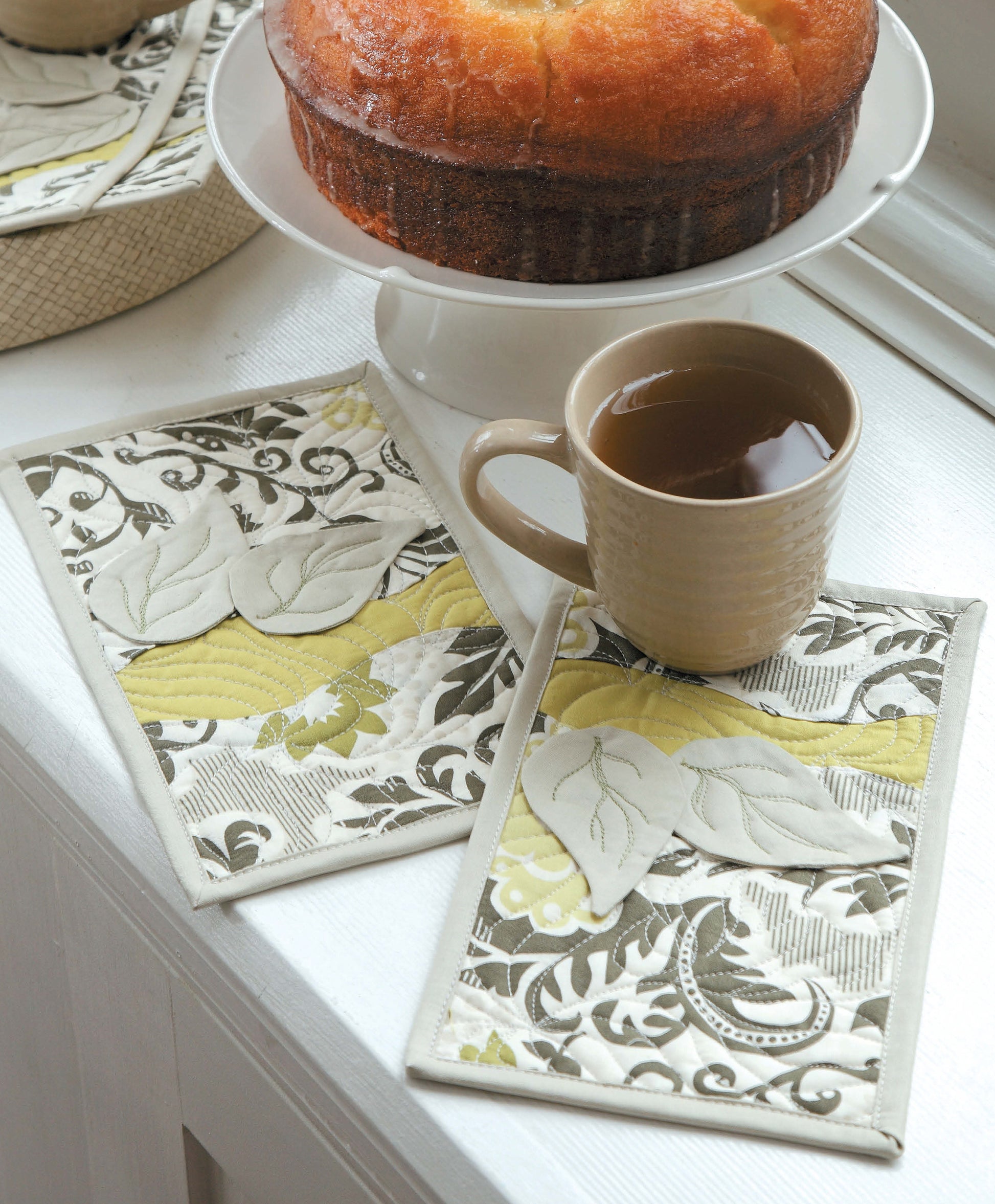 A brown mug filled with tea rests on a Leisure Arts “Learn to Sew Easy Curves” fabric coaster—its pattern echoing the gentle curves featured in the collection—beside a cake on a white stand, with another matching coaster nearby.