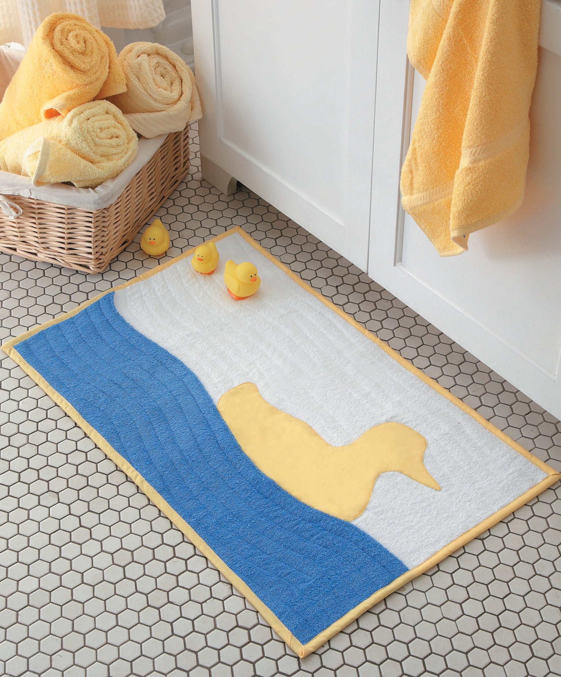 A bath mat shaped like a yellow duck sits on hexagon tile floor, beside three rubber ducks, yellow towels, and a towel basket in a bright bathroom—a cheerful scene that could easily inspire beginner quilt patterns or playful faced applique designs for your next craft project using the Learn to Sew Easy Curves book from Leisure Arts.