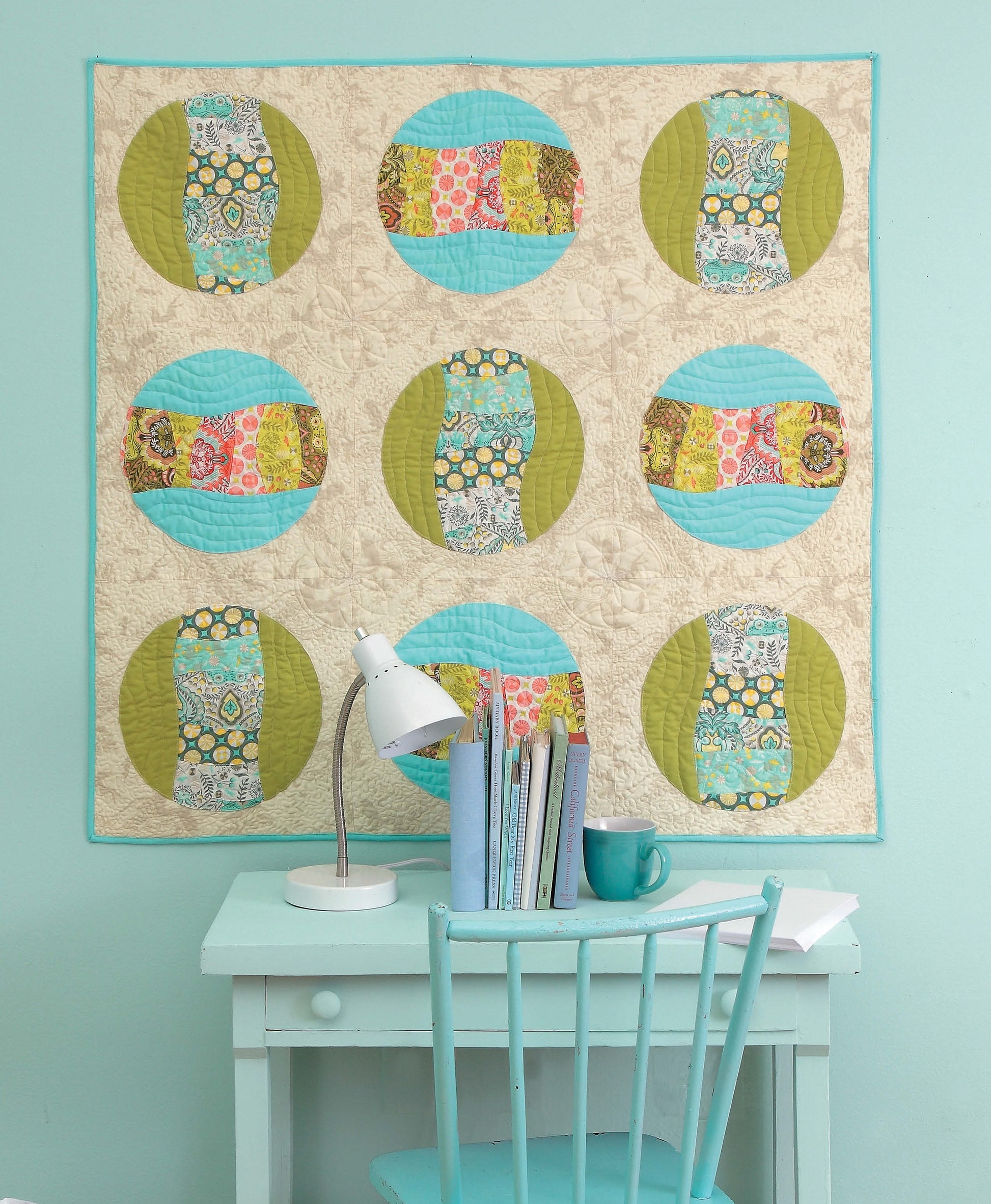Below a light wall, a turquoise desk and chair with books and a lamp sit beneath the colorful “Learn to Sew Easy Curves” quilt by Leisure Arts, which features nine patterned circles and expertly sewn curved seams.