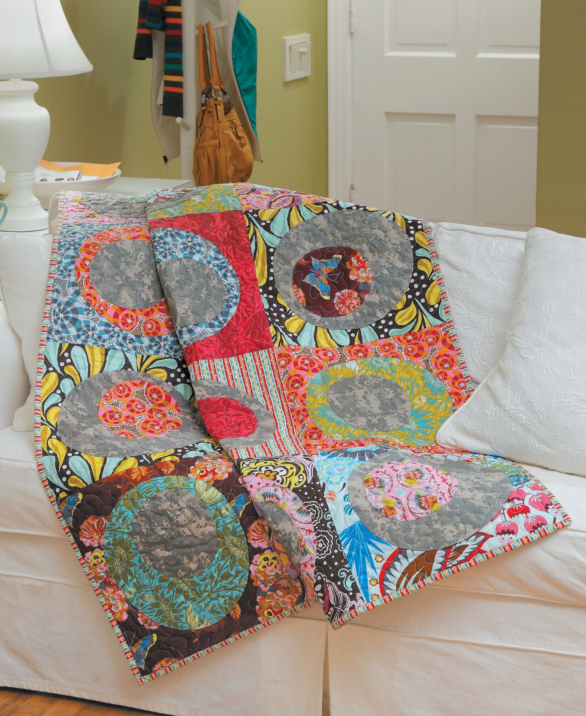 A vibrant quilt created with the “Learn to Sew Easy Curves” pattern by Leisure Arts, showcasing circular patchwork designs and curved seams in assorted bright fabrics, is draped over a white sofa in a cozy living room.