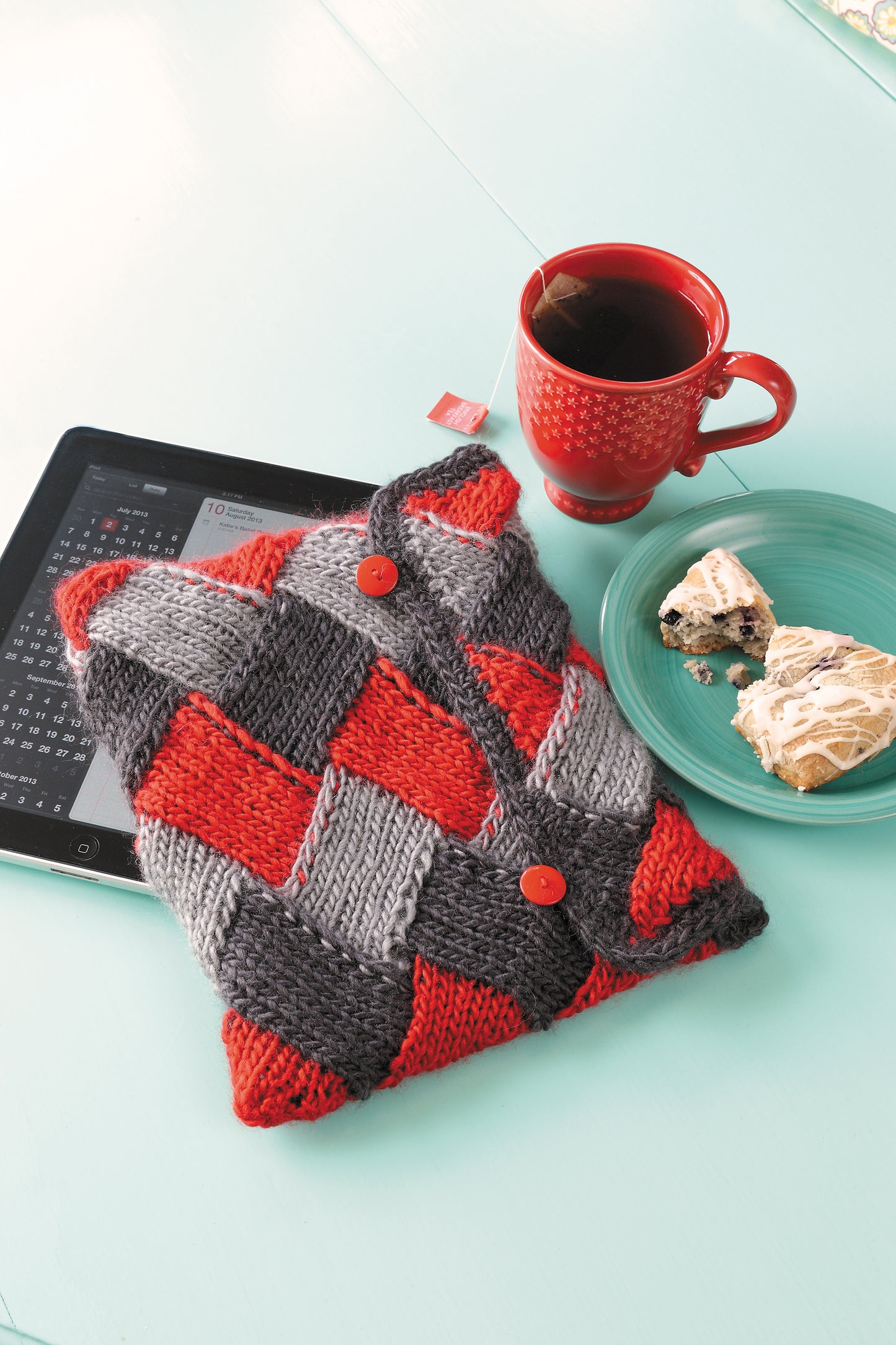 A tablet with "I Can't Believe I'm Entrelac Knitting" by Leisure Arts rests on a turquoise table with a red mug of coffee and a green plate holding a cinnamon roll and scone.