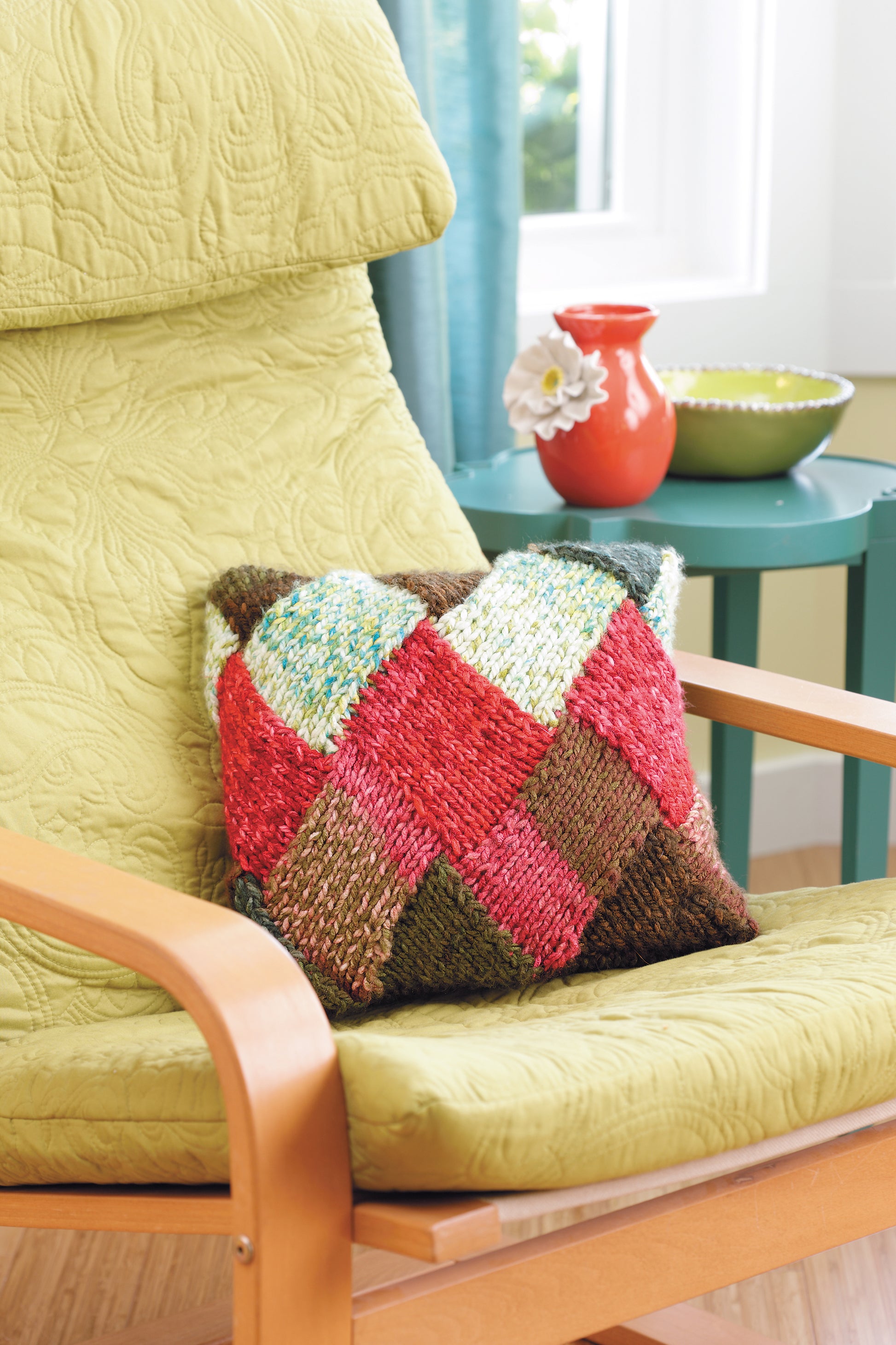 The Leisure Arts "I Can't Believe I'm Entrelac Knitting" digital download features a colorful patchwork pillow for beginner knitters, shown on a light green chair beside a red vase with a white flower and a green bowl, near blue curtains.
