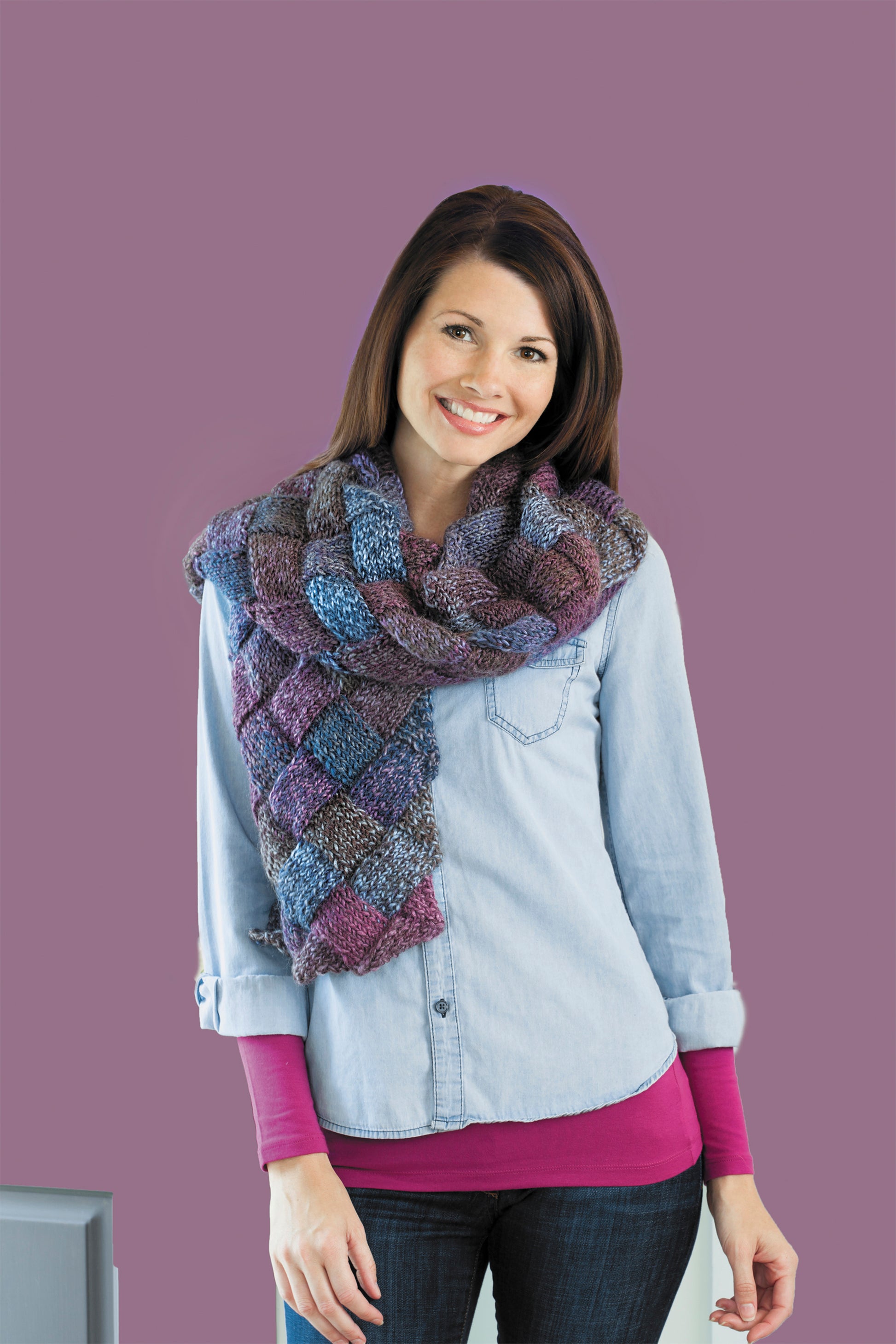 A woman with long brown hair smiles while modeling a large entrelac knit scarf in purple and blue over a light blue shirt, inspired by "I Can't Believe I'm Entrelac Knitting" from Leisure Arts, against a mauve background.