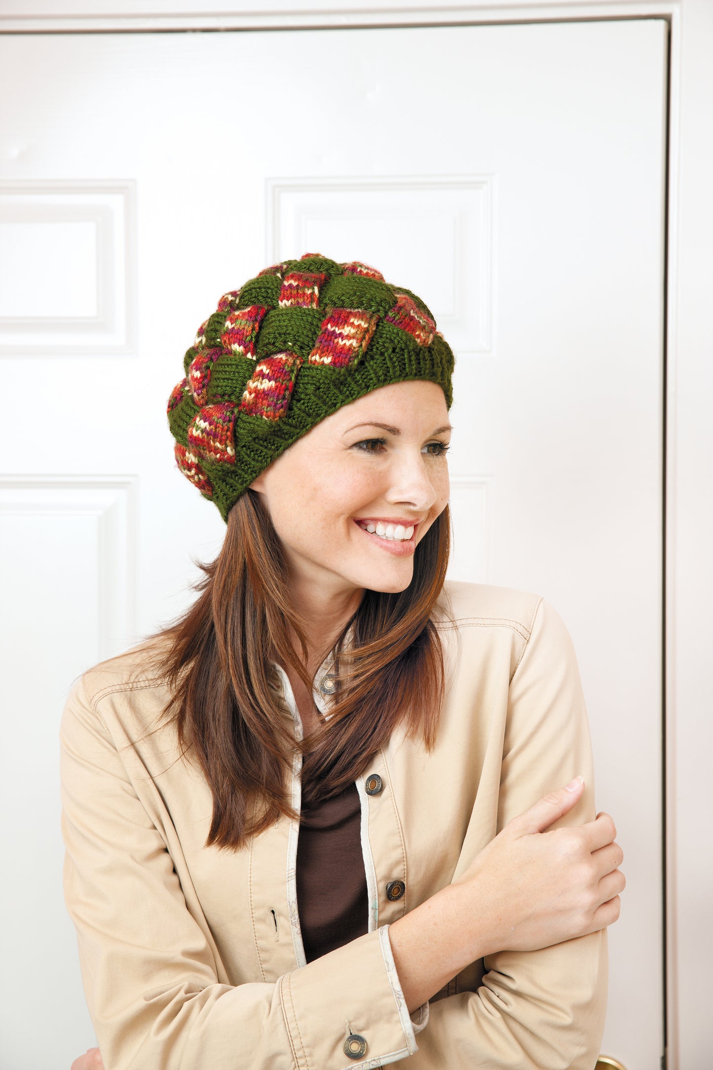 A woman with long brown hair, wearing a beige jacket and a colorful entrelac knit hat—made using "I Can't Believe I'm Entrelac Knitting Digital Download" by Leisure Arts—smiles indoors in front of a white door.