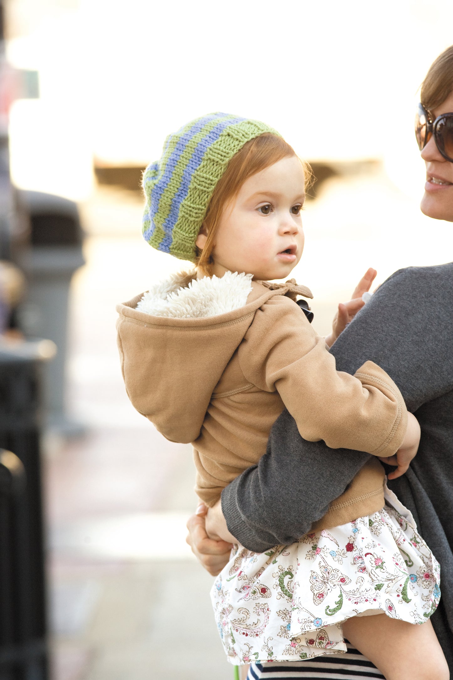 A young child in a green and blue striped hat, possibly crafted with Leisure Arts' "Learn to Knook" digital download, is carried by an adult in a gray sweater. The child looks to the side with curiosity.