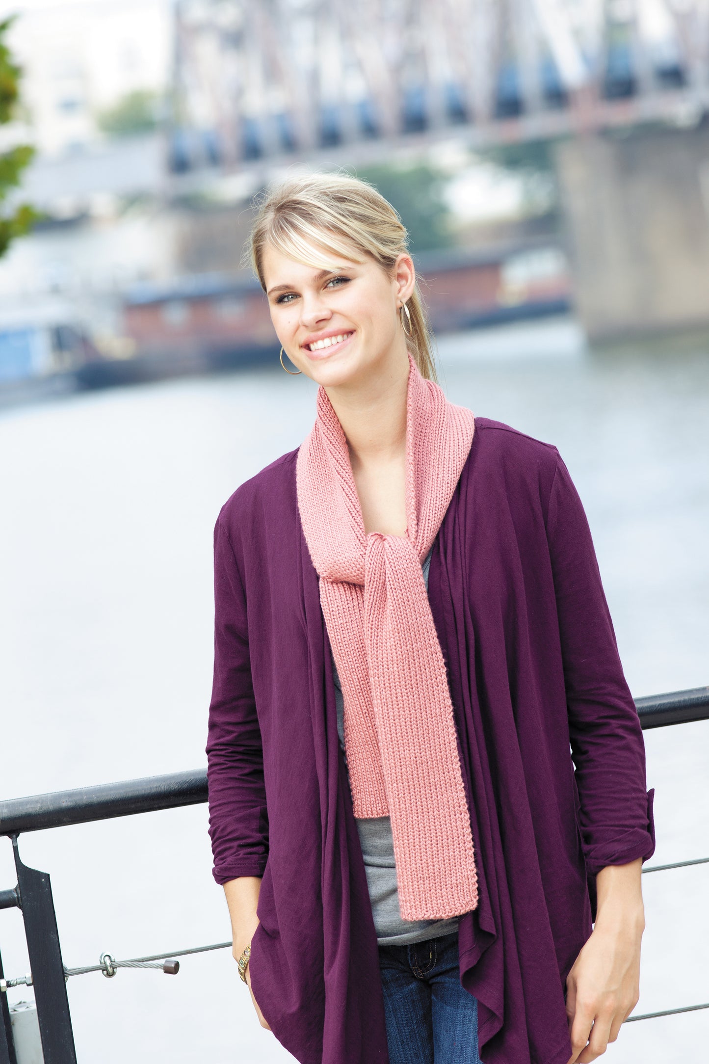 A smiling blonde woman stands by a waterfront railing, wearing a purple cardigan, gray top, and a pink scarf made using the Leisure Arts Learn to Knook beginner crochet kit; a blurred bridge and river appear in the background.