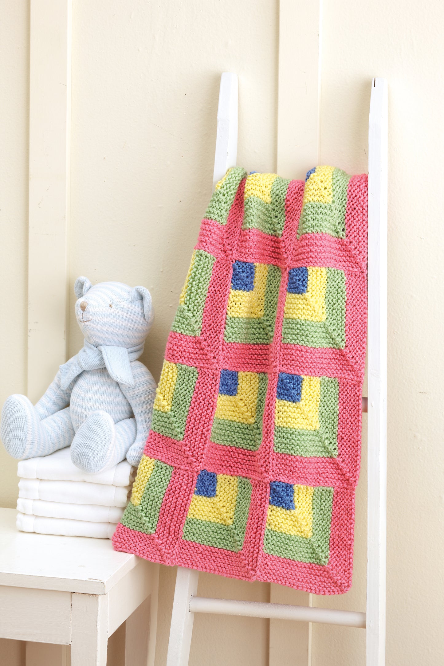 The "Baby Blankets Made with the Knook" by Leisure Arts, featuring charming pastel knit patterns, is draped over a white ladder beside a light blue striped teddy bear and stacked white books against a cream wall.