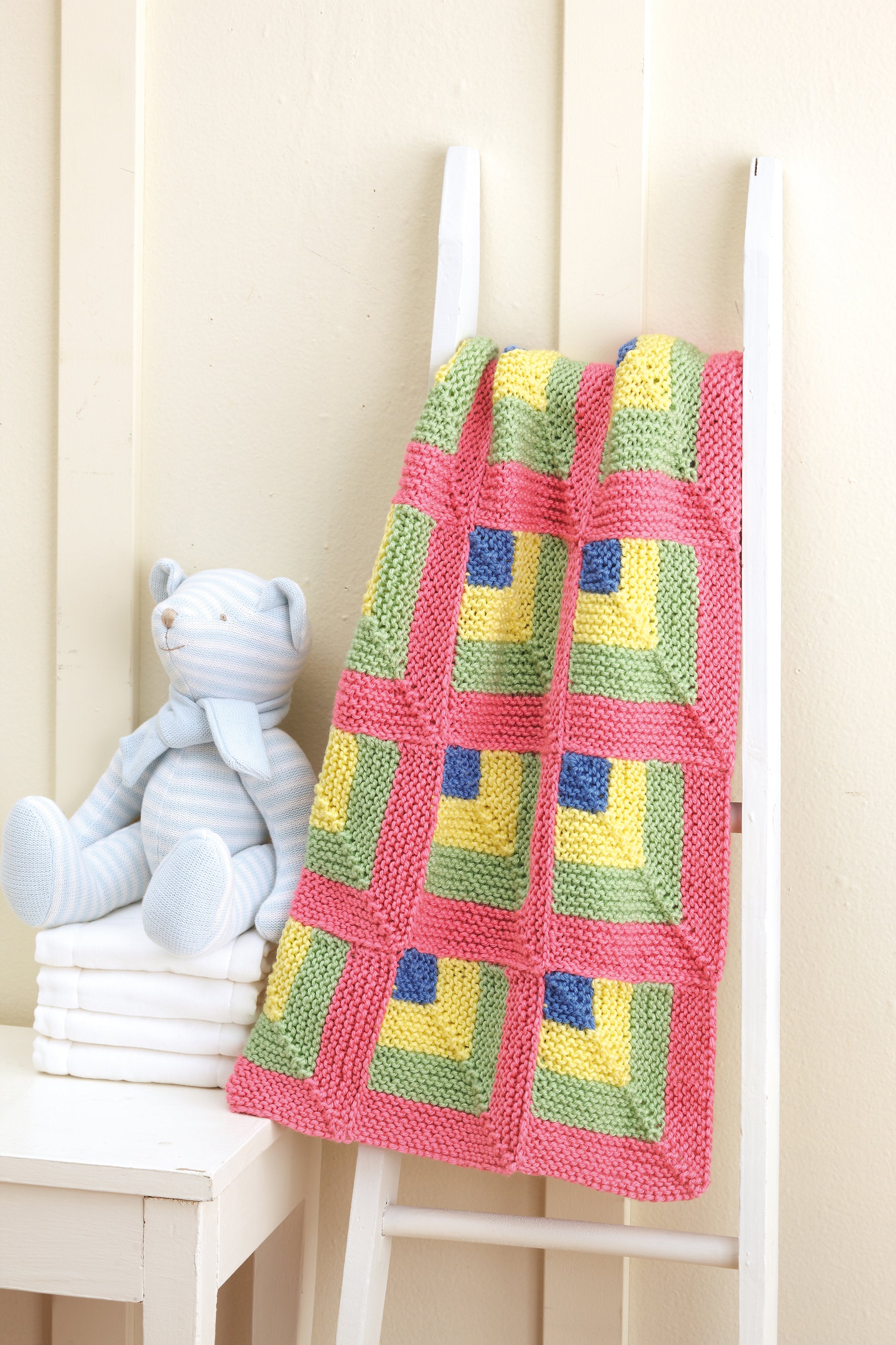 The "Baby Blankets Made with the Knook" by Leisure Arts, featuring charming pastel knit patterns, is draped over a white ladder beside a light blue striped teddy bear and stacked white books against a cream wall.