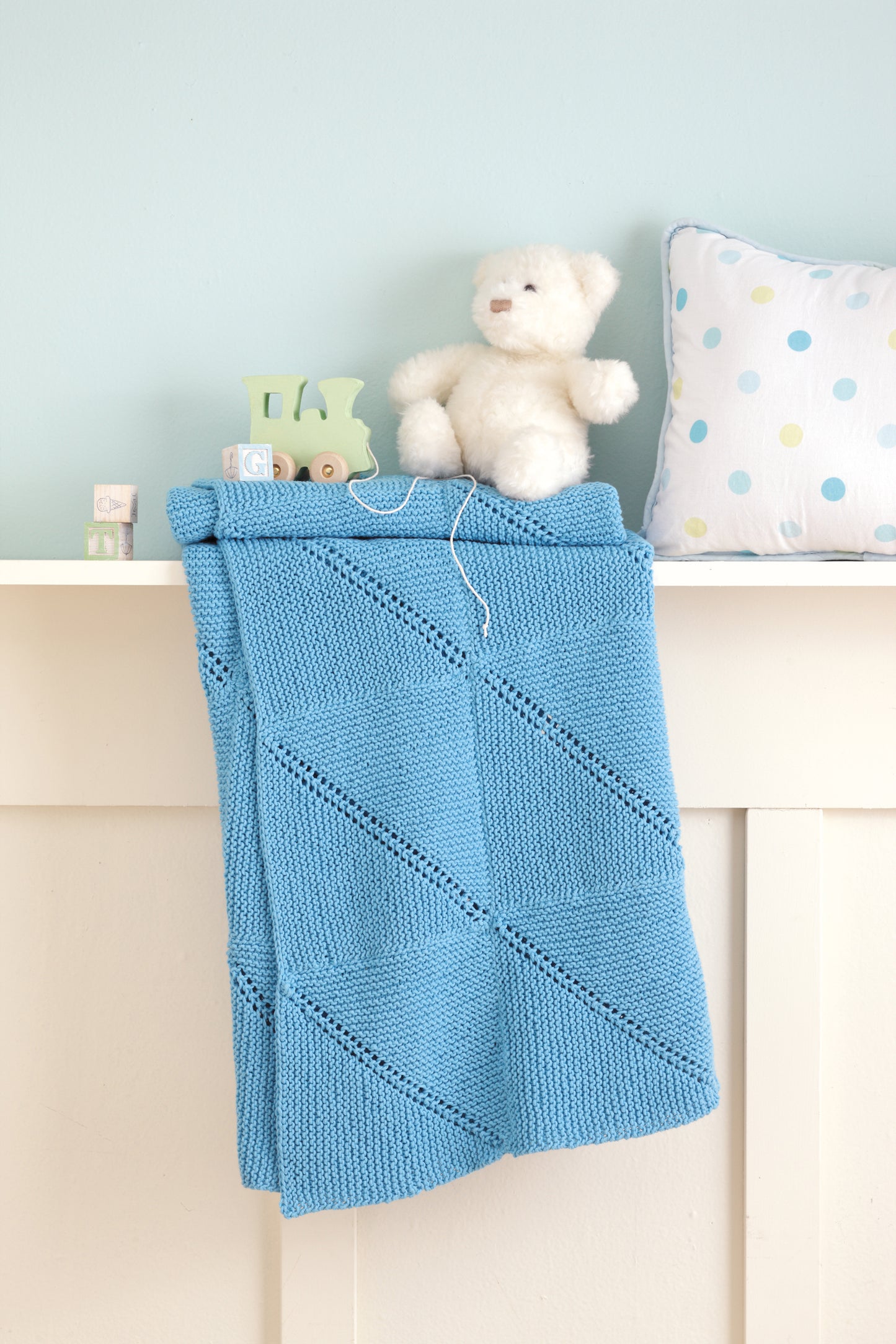 A soft blue "Baby Blankets Made with the Knook" by Leisure Arts hangs over a white shelf holding wooden alphabet blocks, a small white teddy bear, and a pastel polka dot pillow, set against a light blue wall.