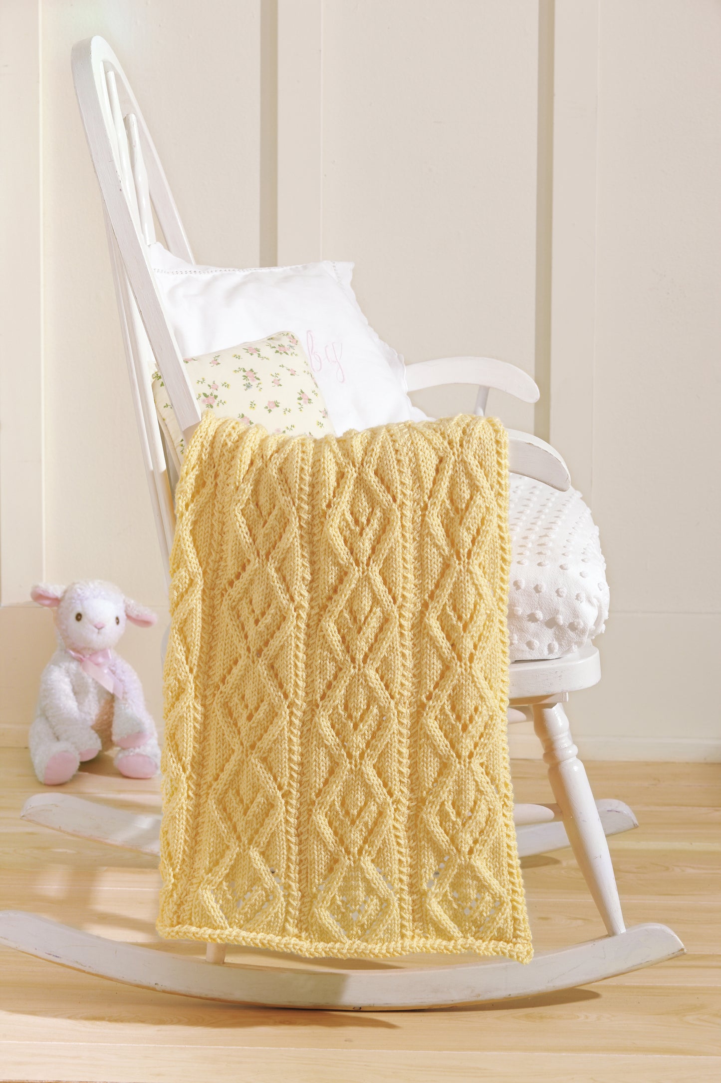 The “Baby Blankets Made with the Knook” by Leisure Arts, in yellow with a diamond pattern, is draped over a white rocking chair beside a white pillow and plush lamb toy on a light wooden floor.