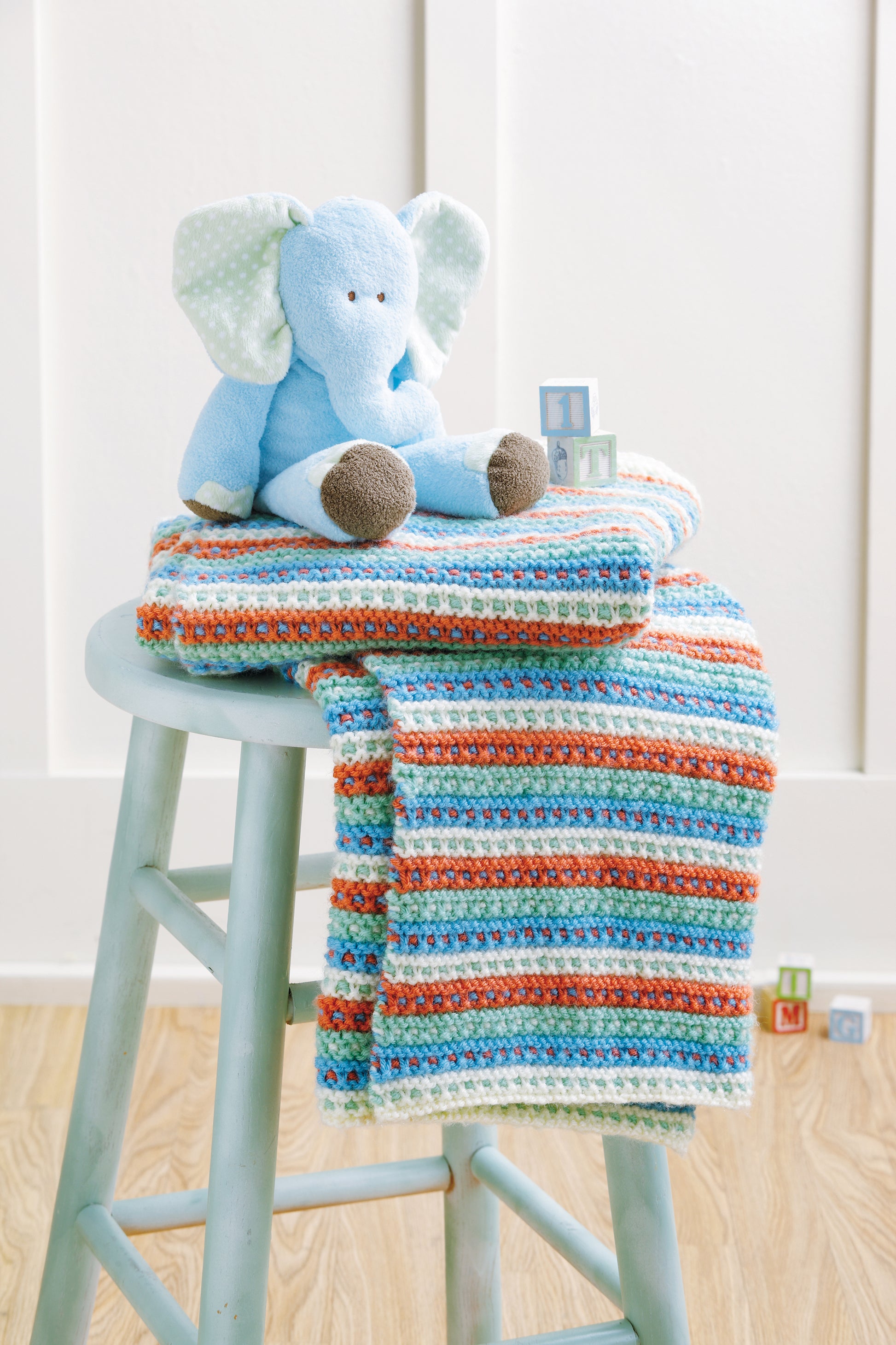 A blue plush elephant sits on a colorful "Baby Blankets Made with the Knook" by Leisure Arts, draped over a light blue stool, while wooden toy blocks are scattered on the floor in the background.