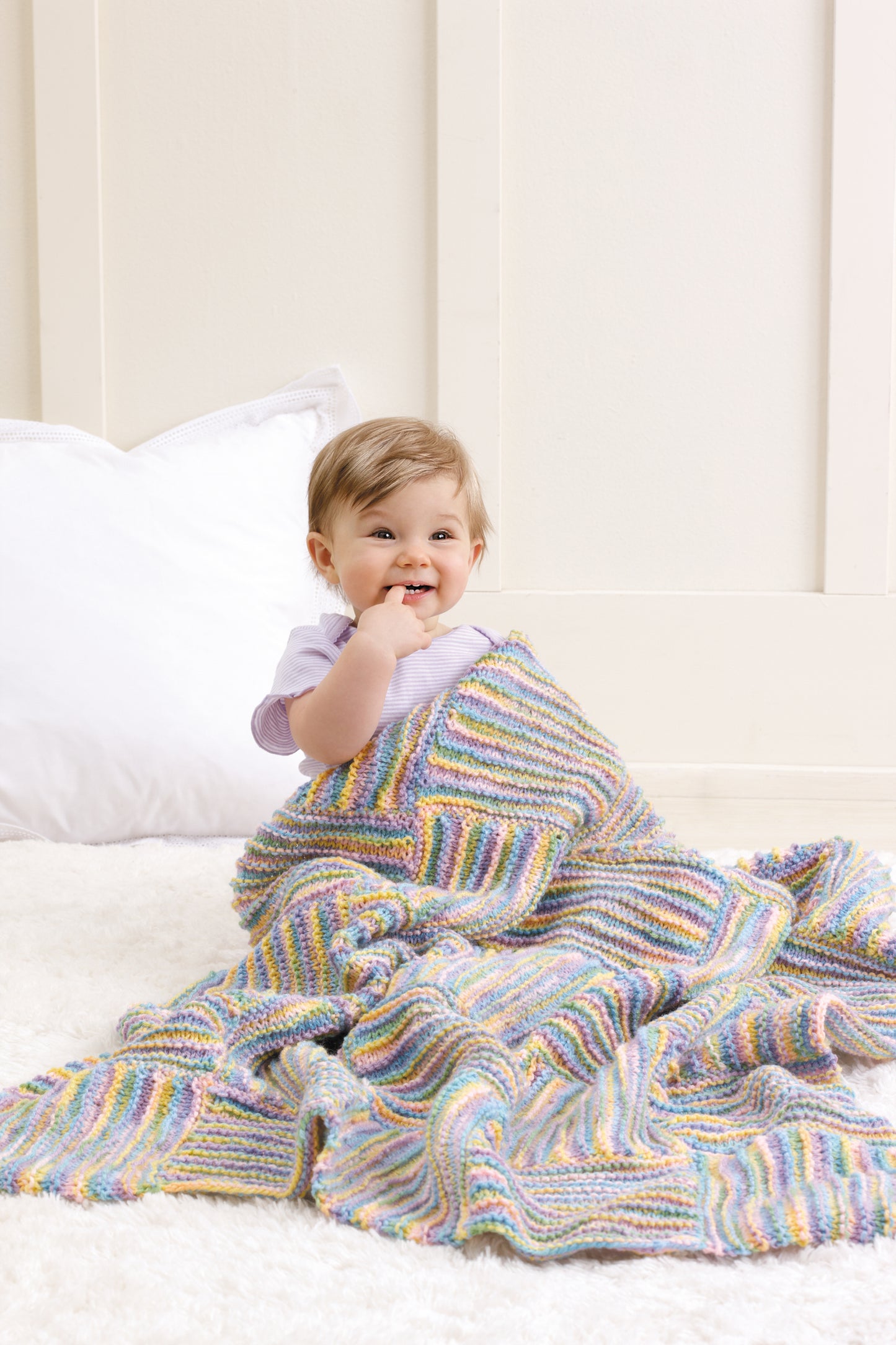 A smiling baby sits on a soft white carpet, wrapped in a colorful blanket from Leisure Arts’ Baby Blankets Made with the Knook, with a white pillow behind and light-colored walls in the background.