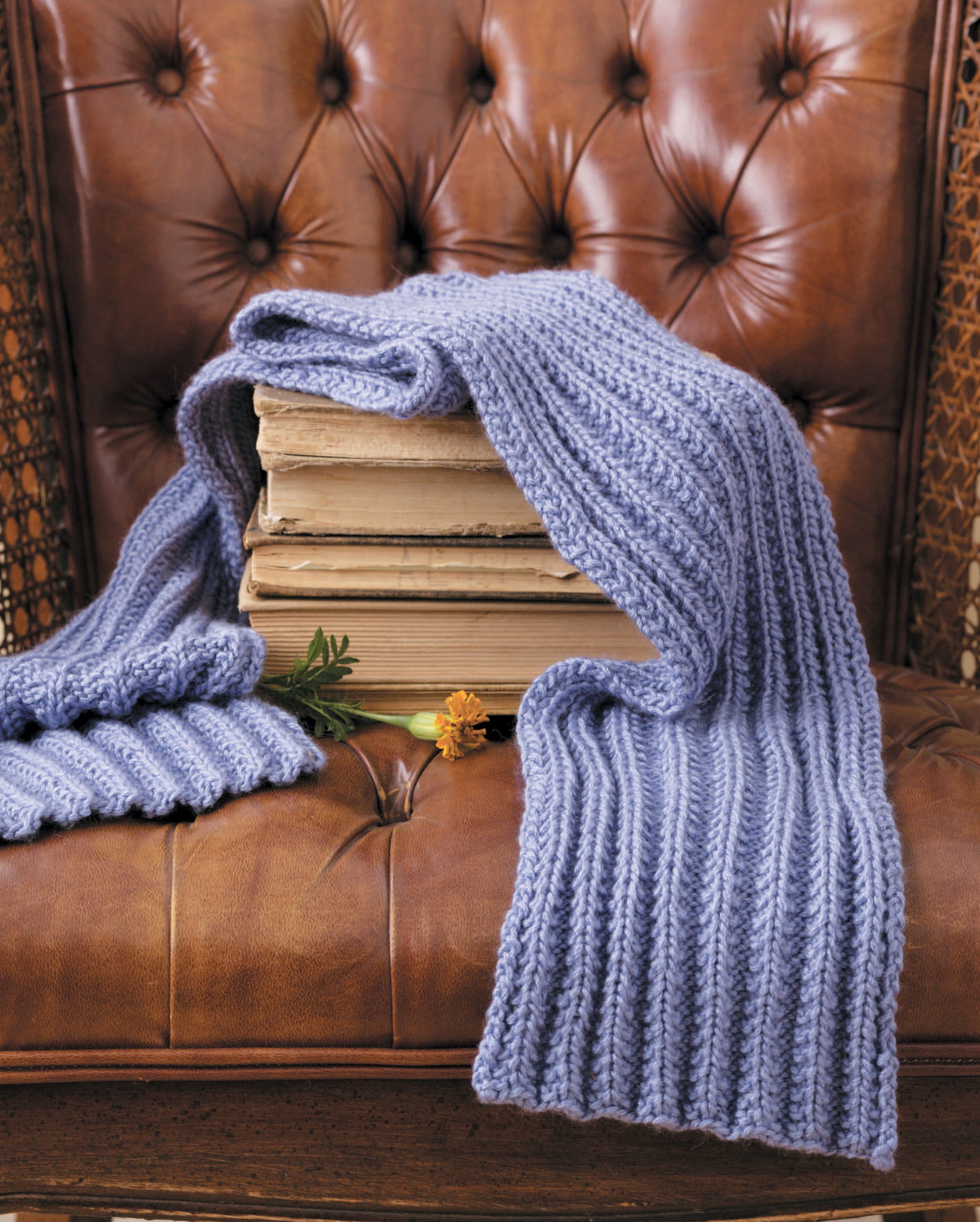 A stack of old books sits on a brown tufted leather chair. Draped over them is a light purple scarf made using “Simple Scarves Made with the Knook” by Leisure Arts, along with two small flowers.