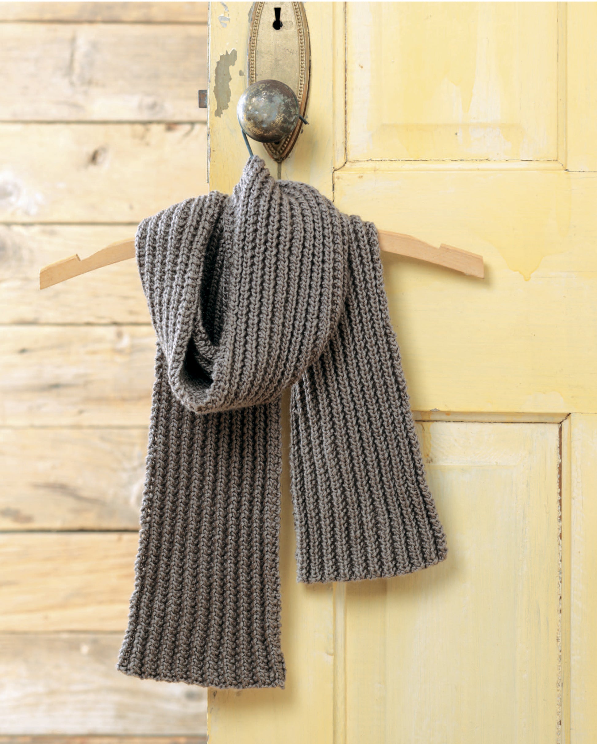 The "Simple Scarves Made with the Knook" by Leisure Arts, a ribbed grey knitted scarf, is displayed on a wooden hanger and round hook against a yellow-painted door with a rustic wood background.