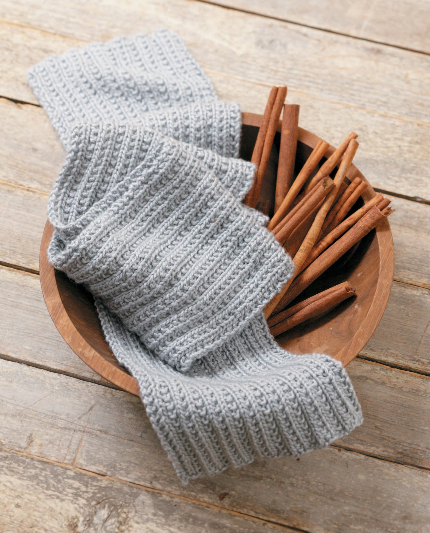 A wooden bowl with cinnamon sticks and a folded, light gray Simple Scarves Made with the Knook by Leisure Arts, crafted in ribbed patterns, placed on a rustic wooden surface.