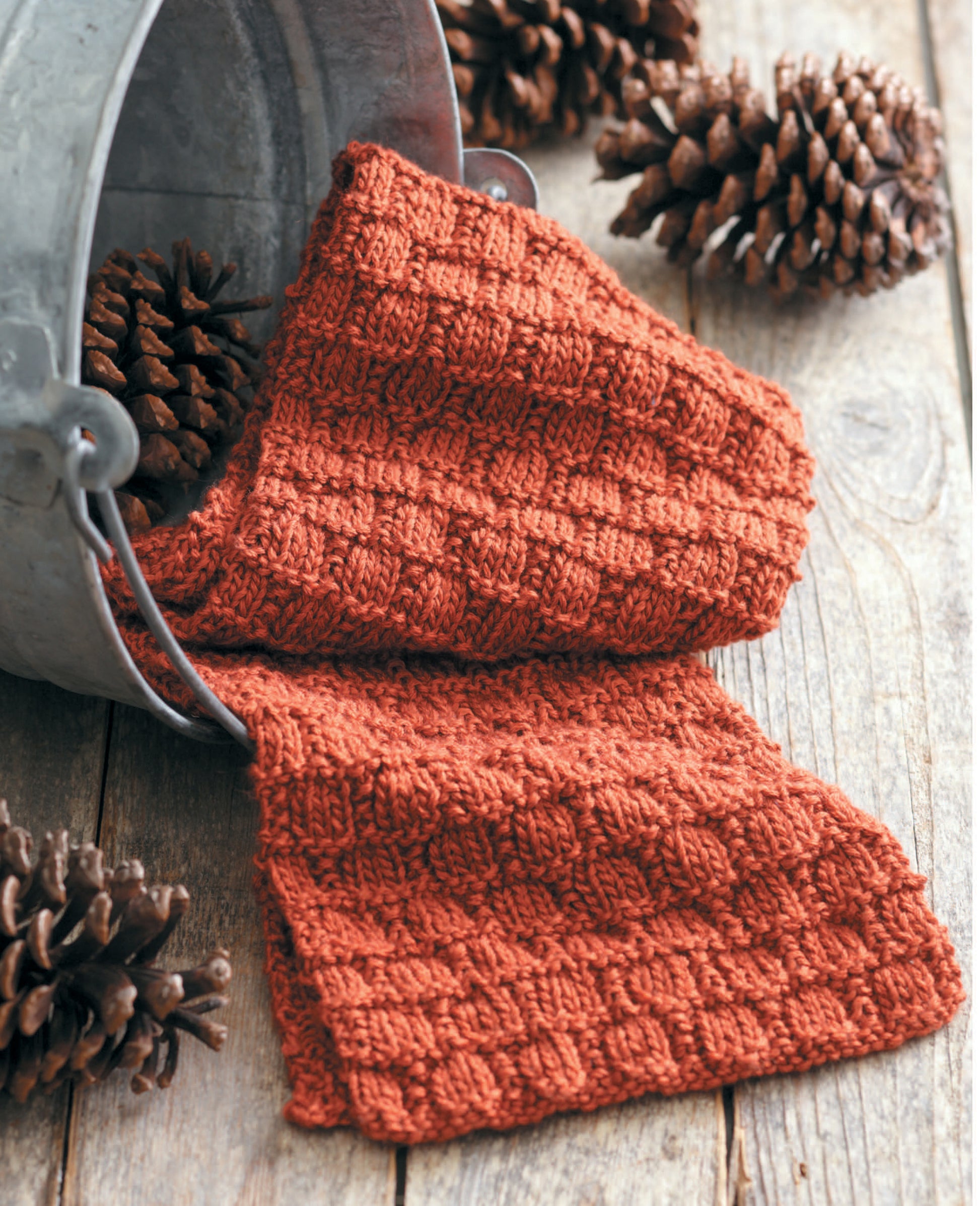 A rust-colored ribbed scarf made with Leisure Arts' "Simple Scarves Made with the Knook" drapes from a metal bucket onto wood, surrounded by pinecones—a timeless pattern for knitters.
