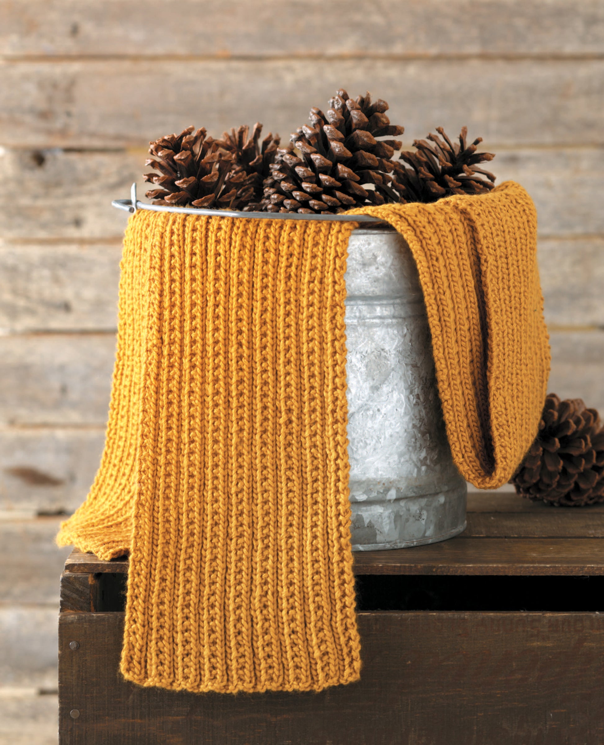 A mustard-yellow scarf from Leisure Arts’ "Simple Scarves Made with the Knook" drapes over a metal bucket of pinecones, set against a rustic wooden background with more pinecones on a nearby dark wooden box.