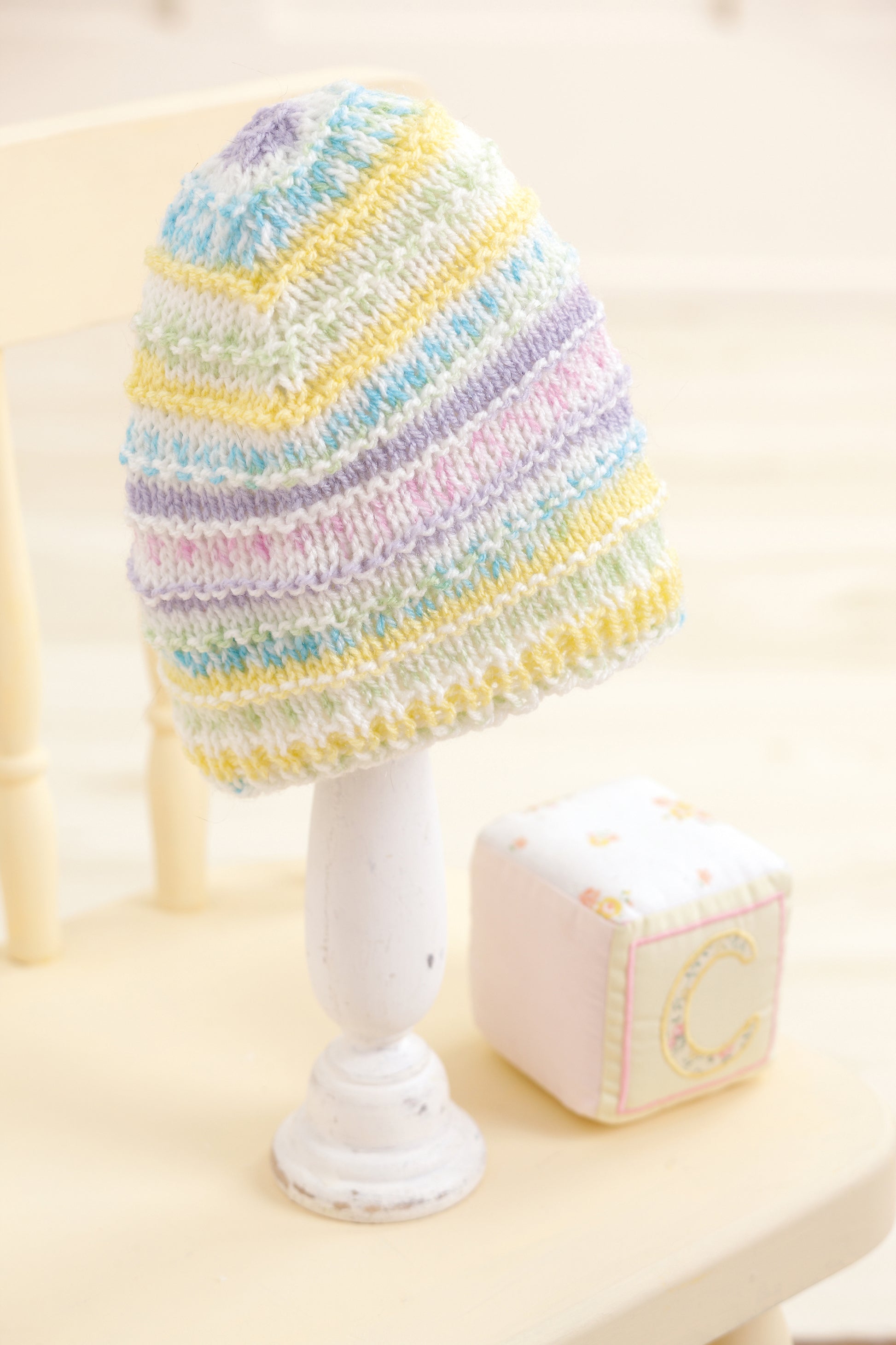 A pastel-striped Baby Beanie Made with the Knook by Leisure Arts is shown on a white wooden stand beside a fabric block with the letter C, all arranged on a pale wooden chair.