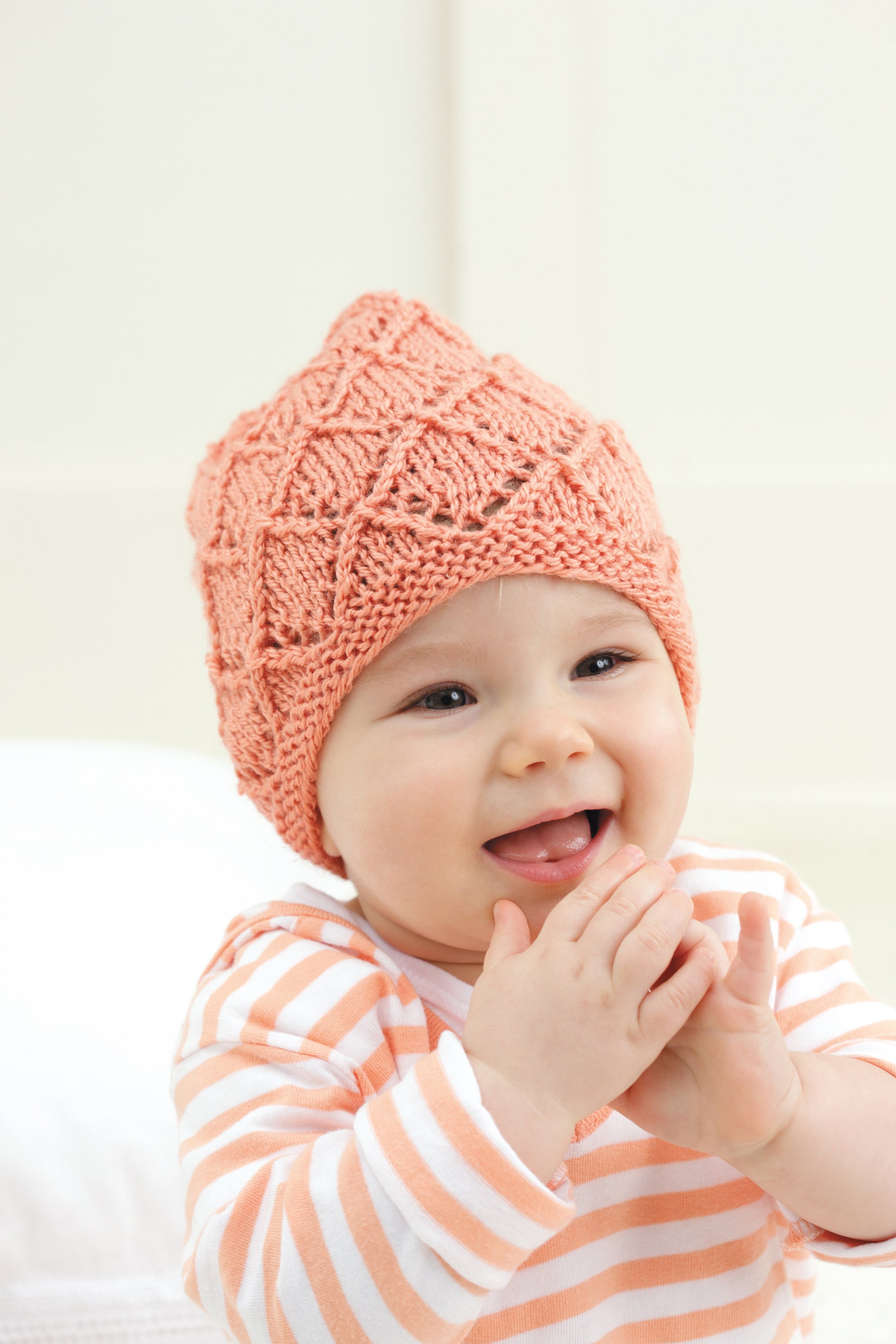 A smiling baby wears a textured, peach-colored "Baby Beanies Made with the Knook" cap by Leisure Arts and a white shirt with orange stripes, holding its hands near its mouth.