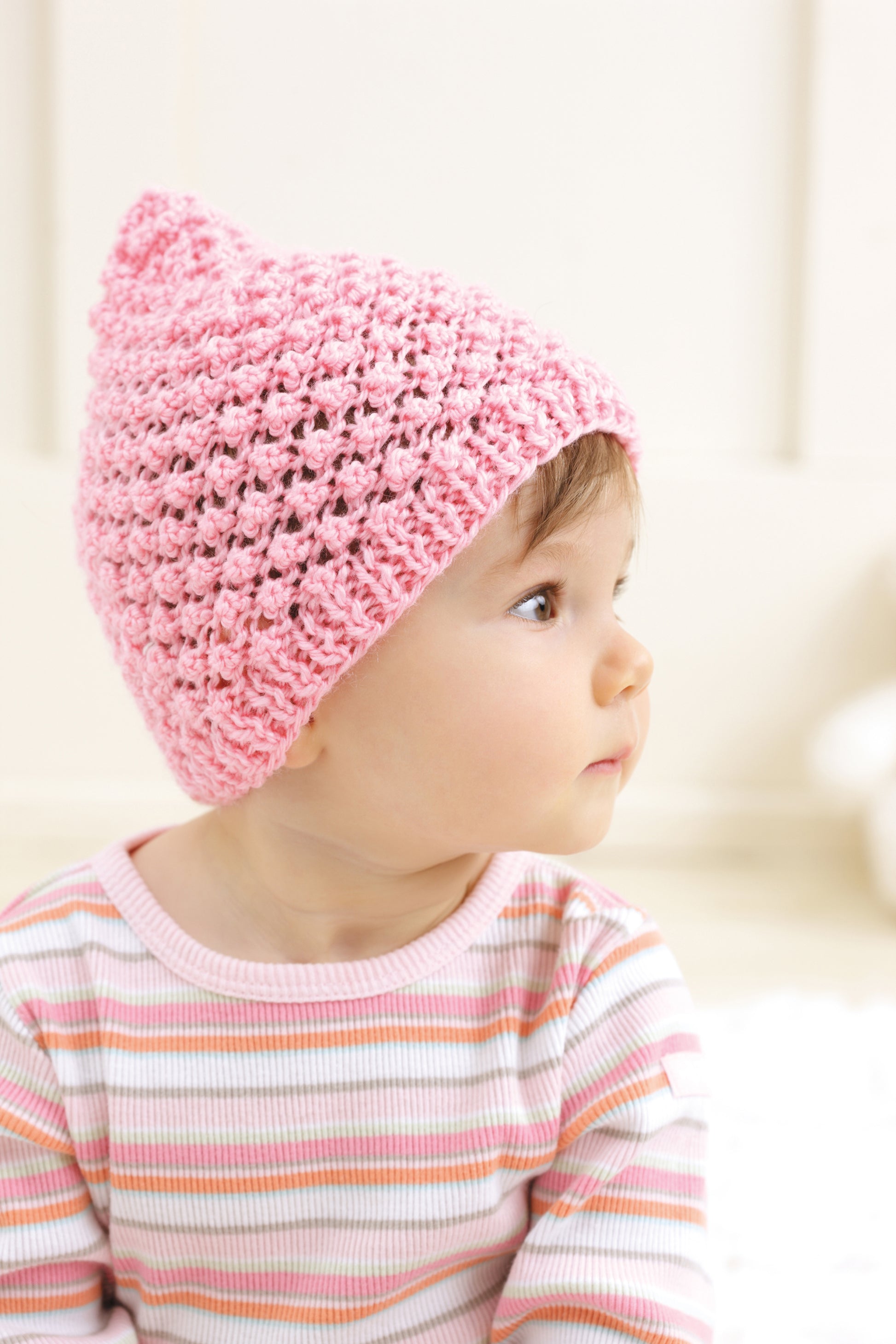 A baby wears a pink beanie from Leisure Arts’ “Baby Beanies Made with the Knook,” paired with a pastel striped shirt. The softly blurred background gives this cozy baby cap image a warm, gentle feel.