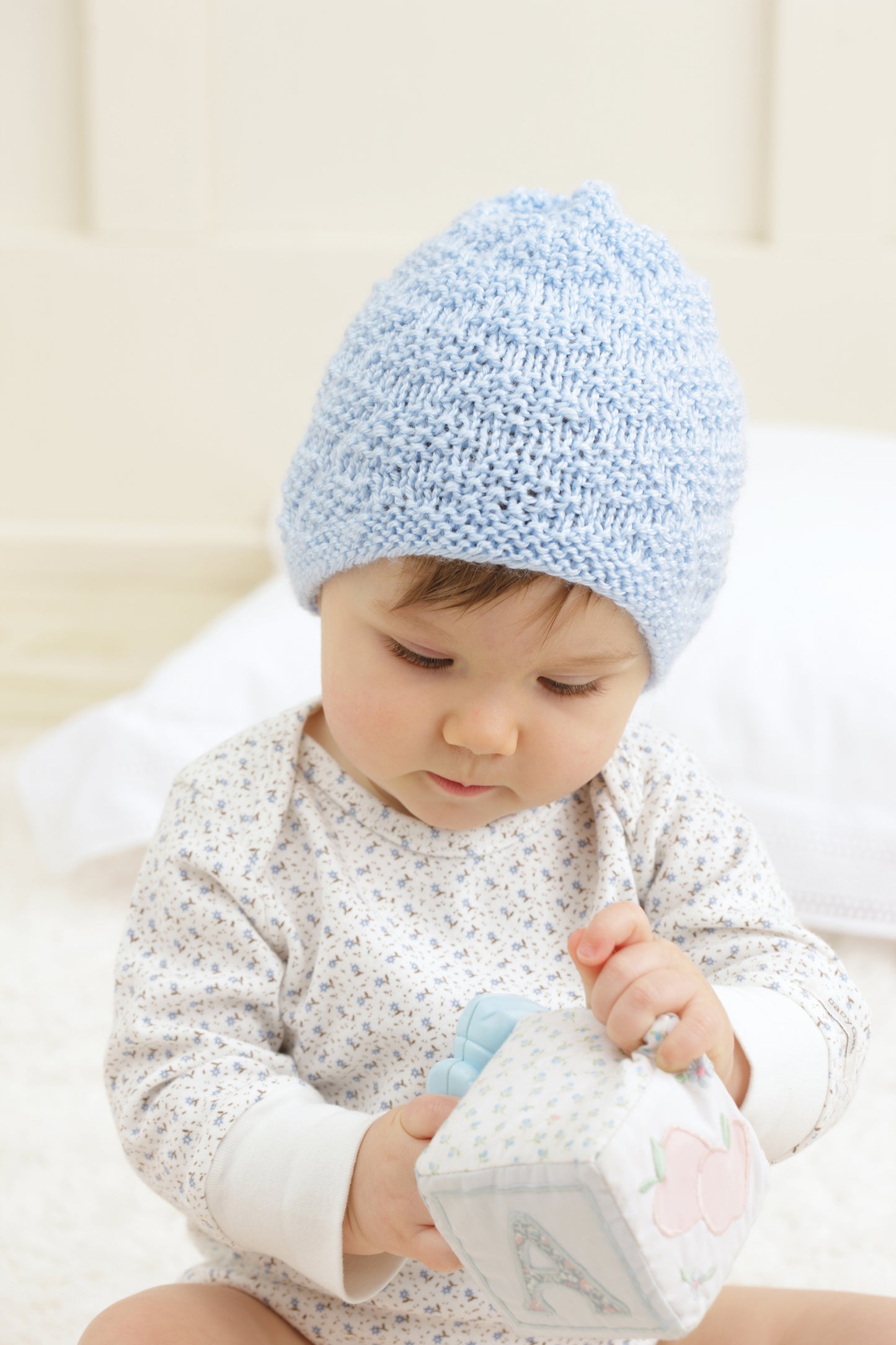 A baby wearing a light blue beanie from "Baby Beanies Made with the Knook" by Leisure Arts sits on a soft surface and plays with a fabric block.