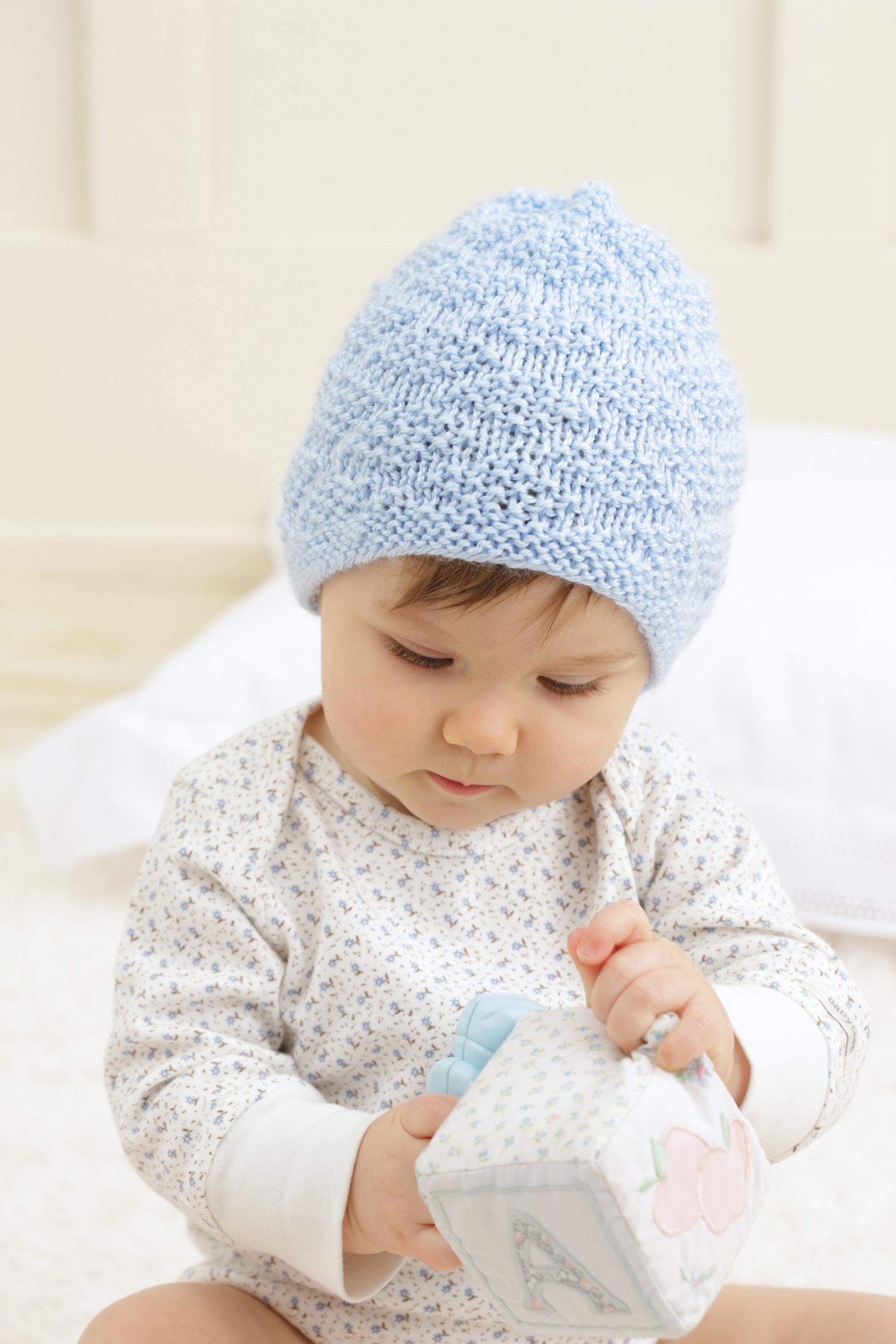 A baby wearing a light blue beanie from "Baby Beanies Made with the Knook" by Leisure Arts sits on a soft surface and plays with a fabric block.