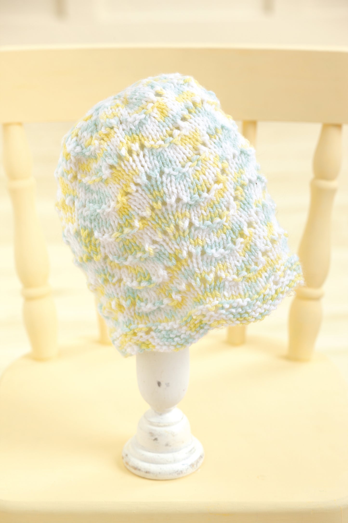 The Leisure Arts "Baby Beanies Made with the Knook" is a pastel-colored knitted baby cap in soft blue, yellow, and white hues, shown on a white stand atop a light wooden chair. It features a textured pattern and cozy fabric for comfort.