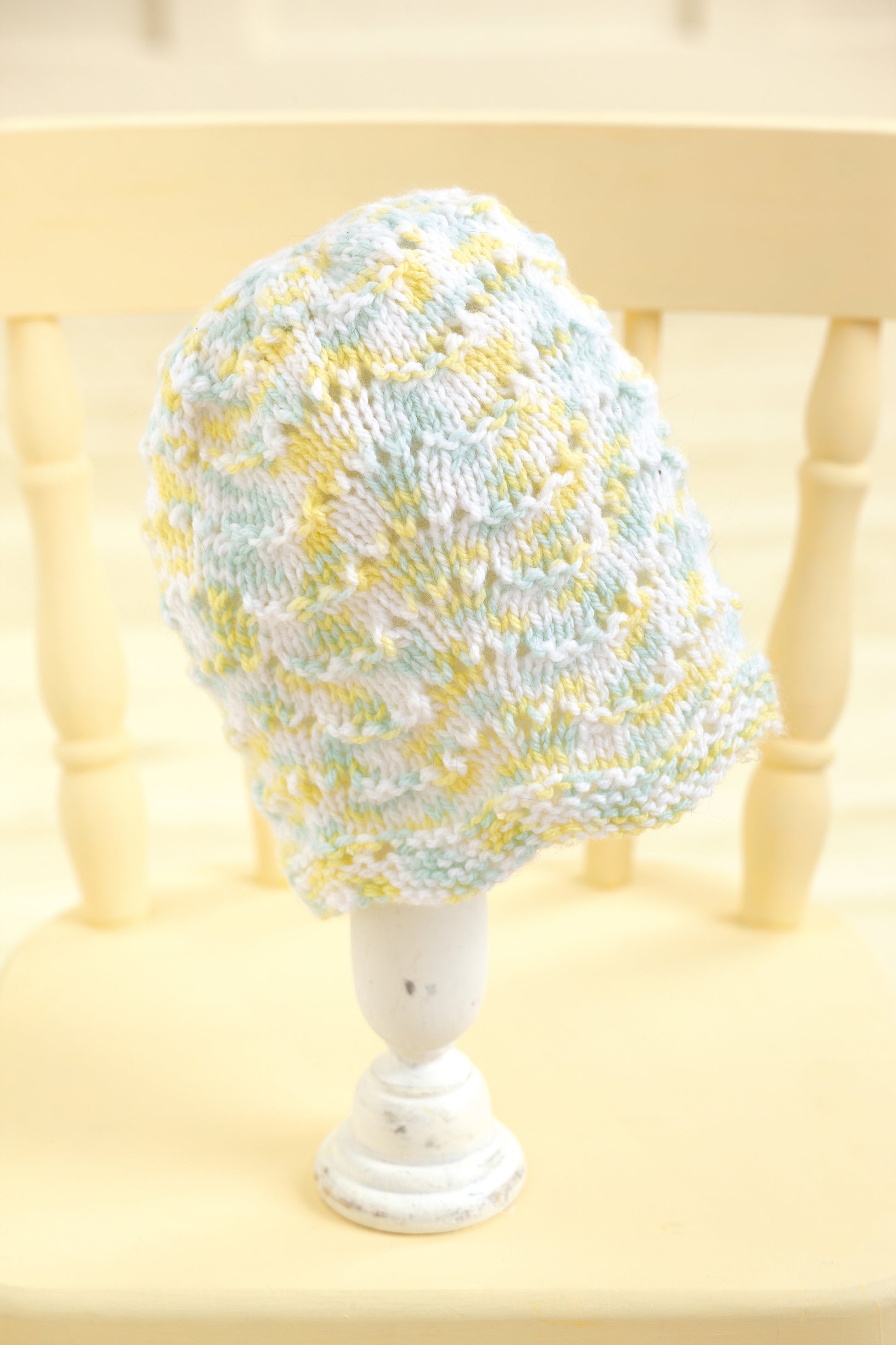 The Leisure Arts "Baby Beanies Made with the Knook" is a pastel-colored knitted baby cap in soft blue, yellow, and white hues, shown on a white stand atop a light wooden chair. It features a textured pattern and cozy fabric for comfort.