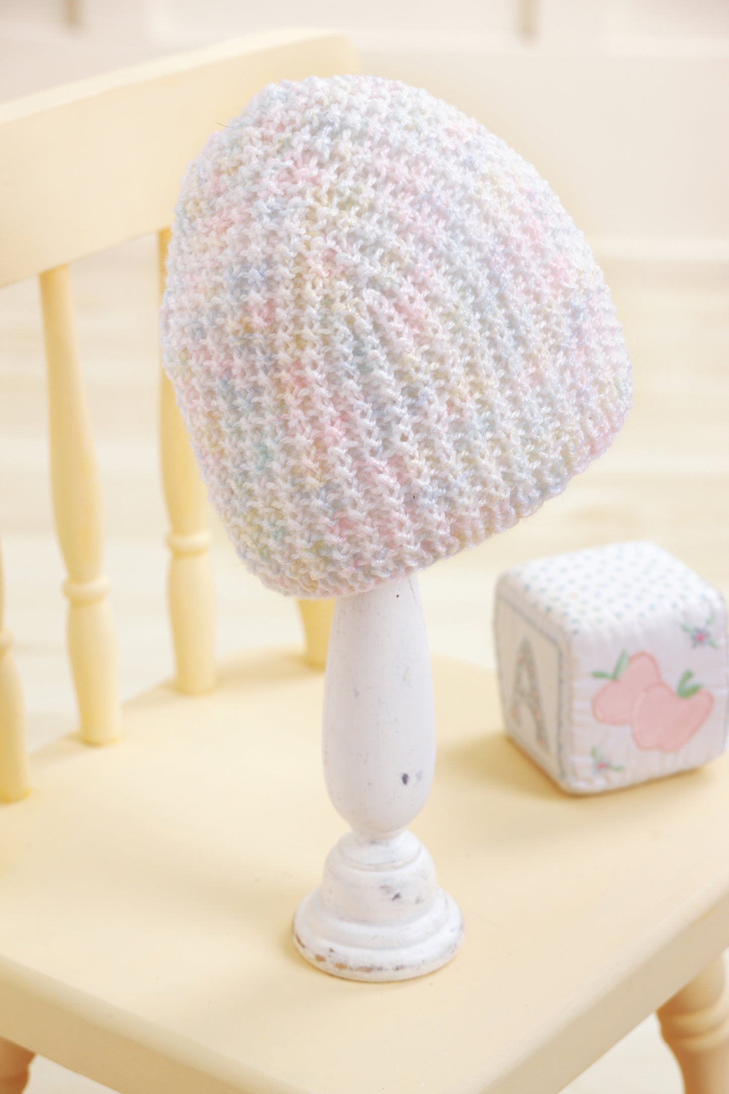A pastel-colored "Baby Beanies Made with the Knook" by Leisure Arts is displayed on a white stand atop a pale yellow chair, with a soft block showing an embroidered apple and the letter “A” in the background.