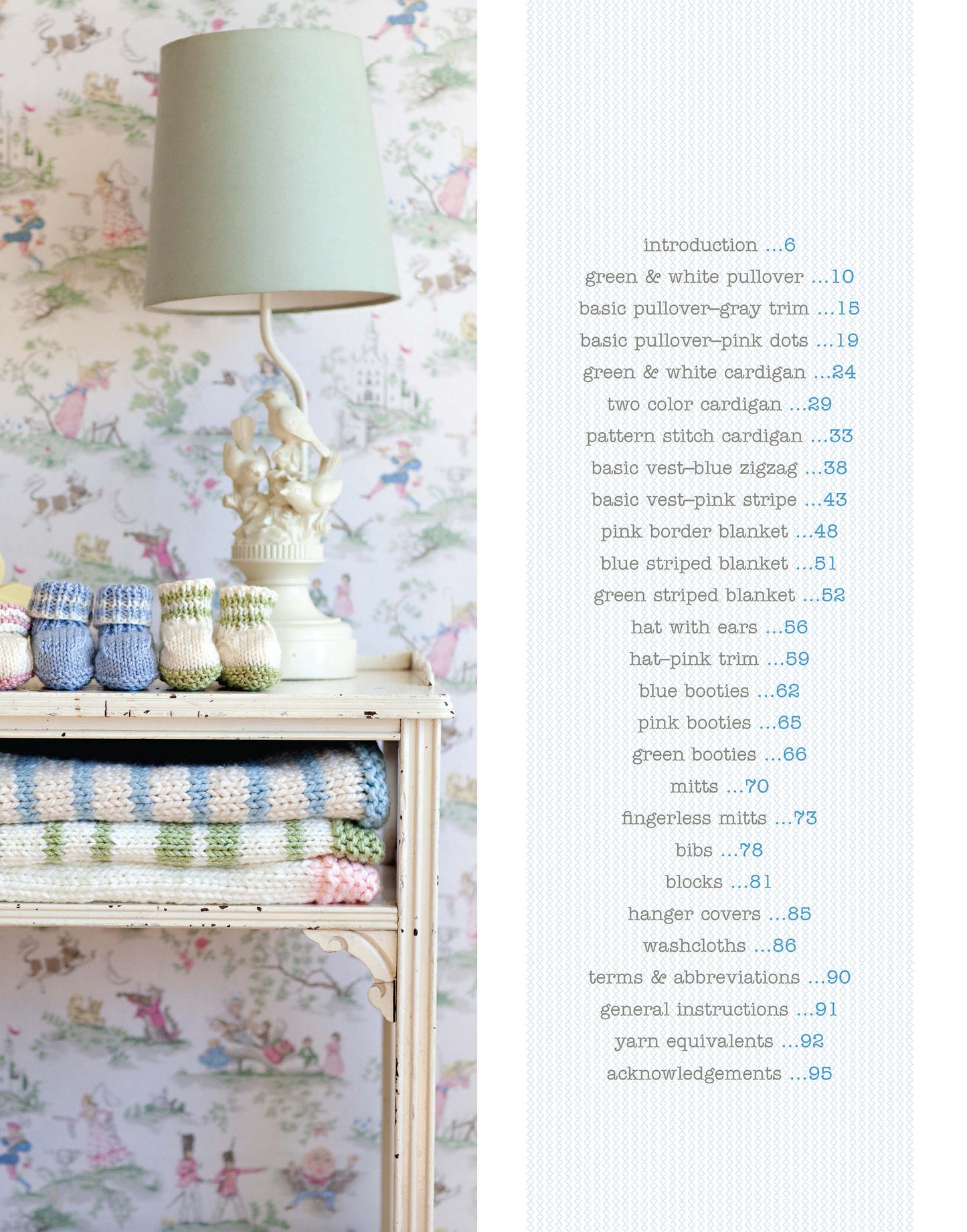 Against floral wallpaper, a vintage table displays pastel knitted baby items, folded blankets, and a sage lamp. Inside "Knit in a Day for Baby" by Leisure Arts, find quick and easy baby knitting projects—ideal for last-minute gifts.