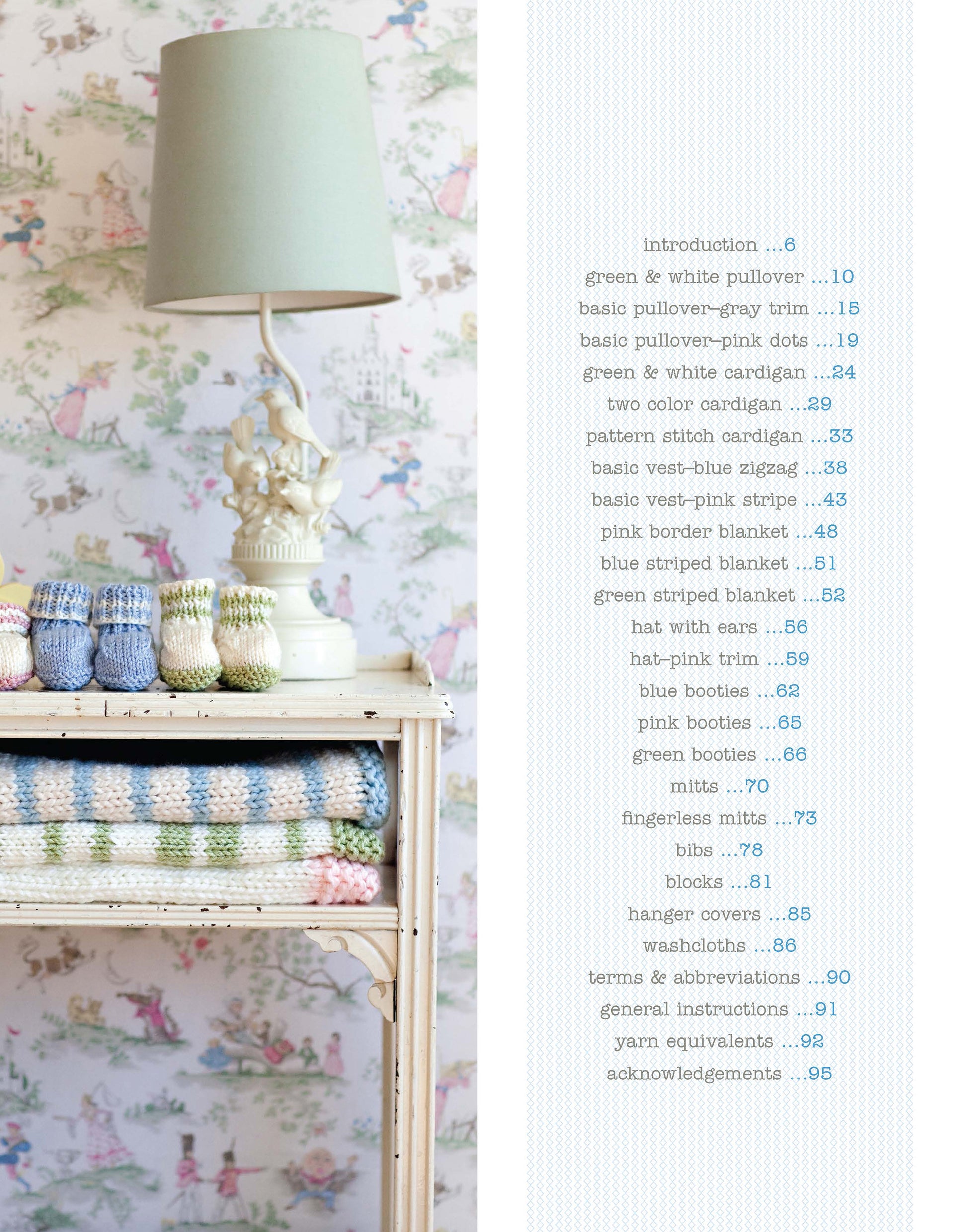 Against floral wallpaper, a vintage table displays pastel knitted baby items, folded blankets, and a sage lamp. Inside "Knit in a Day for Baby" by Leisure Arts, find quick and easy baby knitting projects—ideal for last-minute gifts.