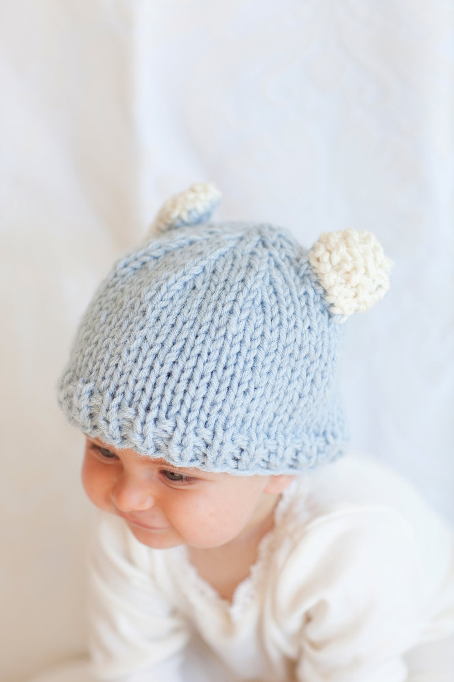 A baby in a light blue knitted hat with cream bear ears—made from Leisure Arts' "Knit in a Day for Baby"—smiles while looking down, dressed in white against a soft, light background.
