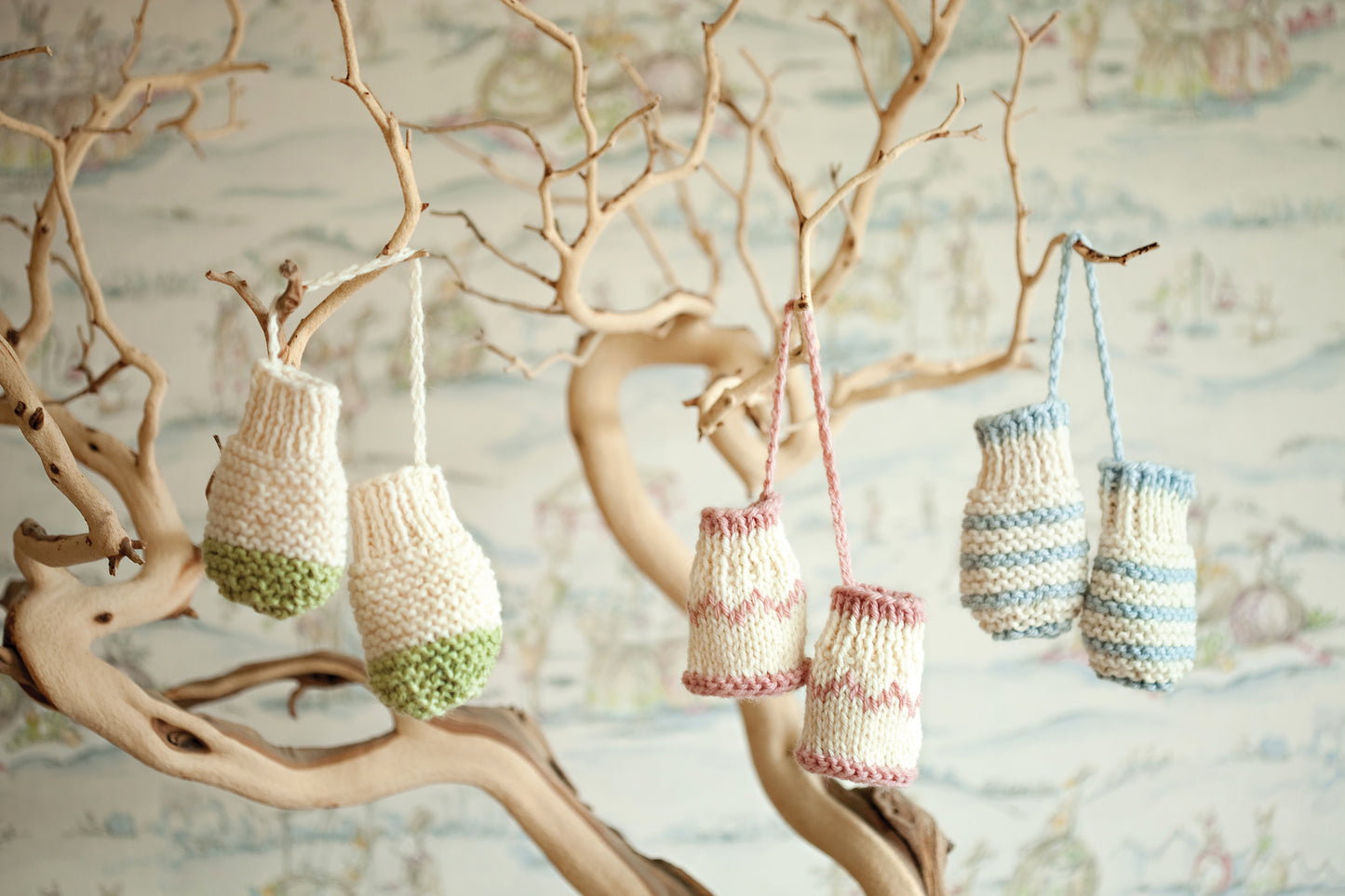 Hand-knitted miniature mittens, inspired by Leisure Arts’ "Knit in a Day for Baby," hang from a bare, twisted tree against a backdrop of softly blurred, pastel-patterned wallpaper.