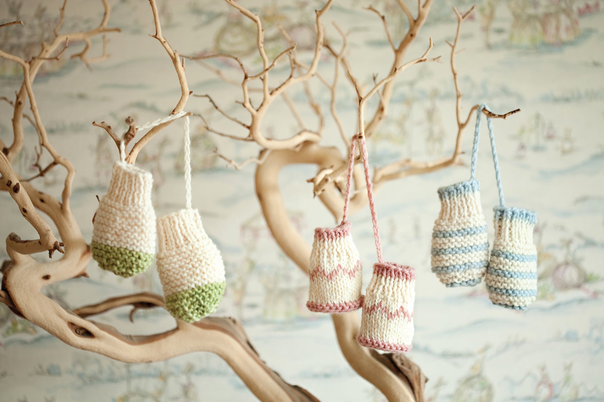 Hand-knitted miniature mittens, inspired by Leisure Arts’ "Knit in a Day for Baby," hang from a bare, twisted tree against a backdrop of softly blurred, pastel-patterned wallpaper.
