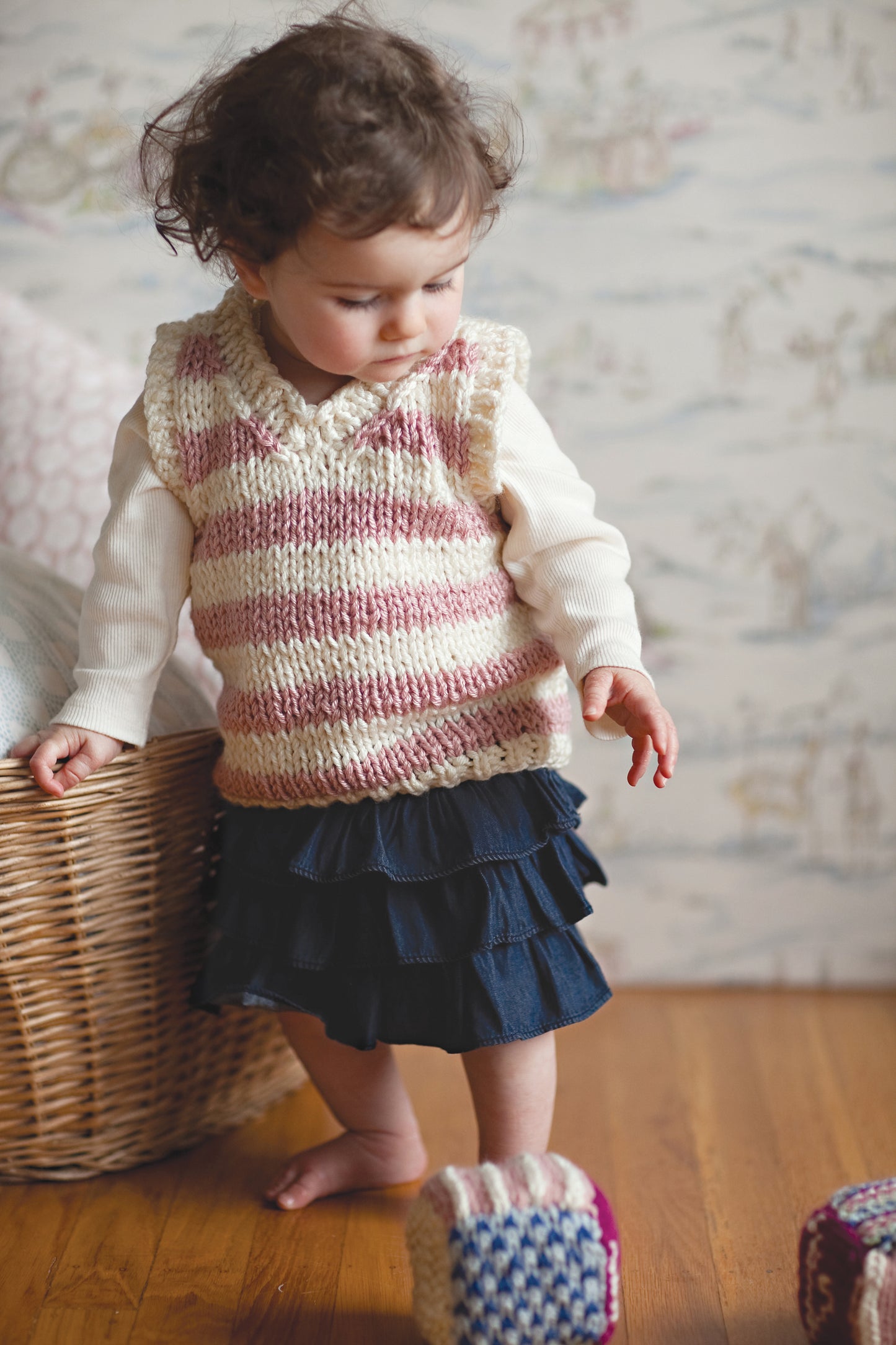 A toddler with curly hair wears a pink and white striped vest made from Knit in a Day for Baby by Leisure Arts, paired with a white long-sleeve shirt and navy skirt, standing on a wooden floor near a basket and colorful blocks.