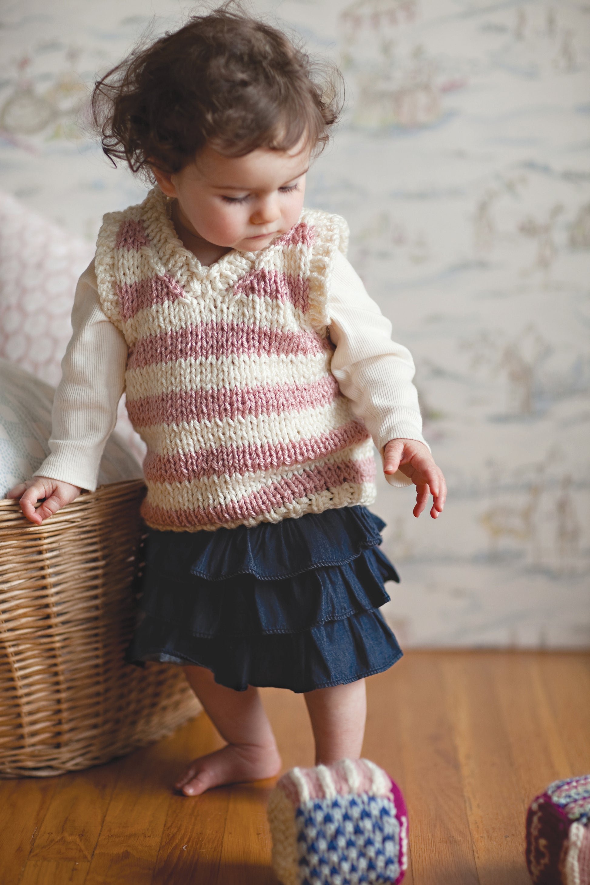 A toddler with curly hair wears a pink and white striped vest made from Knit in a Day for Baby by Leisure Arts, paired with a white long-sleeve shirt and navy skirt, standing on a wooden floor near a basket and colorful blocks.