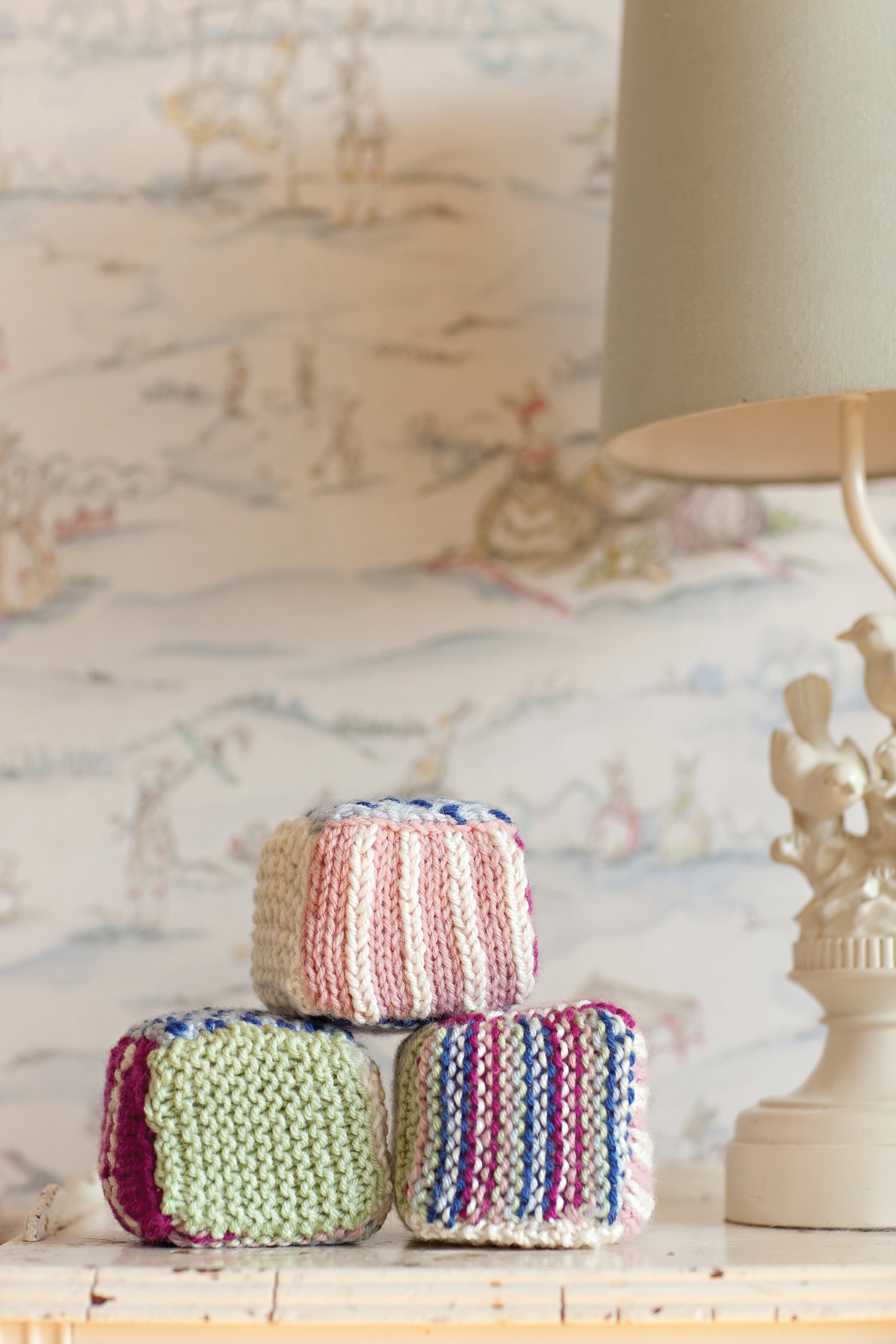 Three colorful blocks from Leisure Arts’ “Knit in a Day for Baby” are hand-knitted and stacked near a lamp with bird figures, set before playful wallpaper with pastel designs.