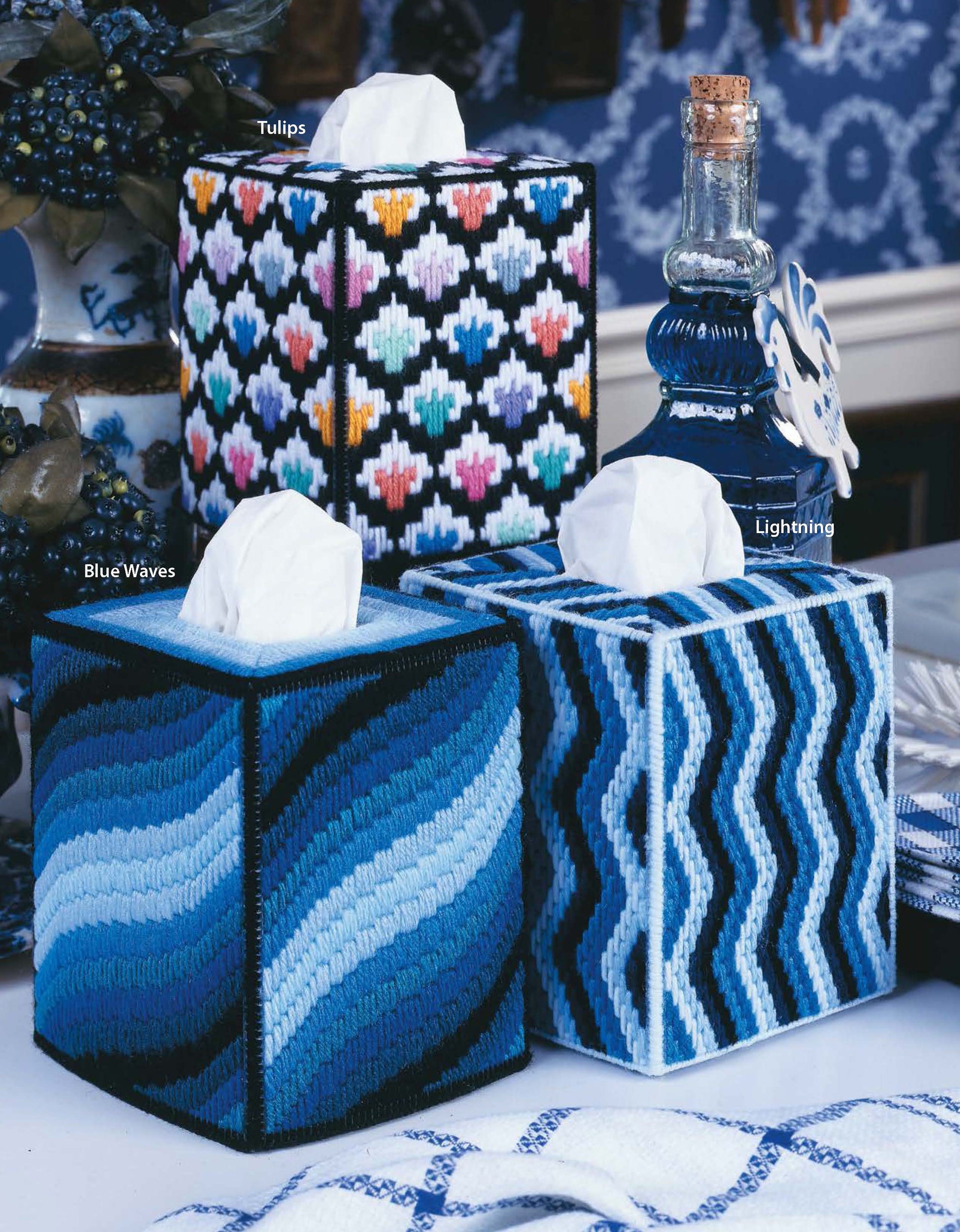 Three Top-Notch Tissue Box Covers by Leisure Arts sit on a table: tulip, blue waves, and blue-white zigzag designs. Blue decor and a glass bottle in the background—perfect beginner craft digital download.