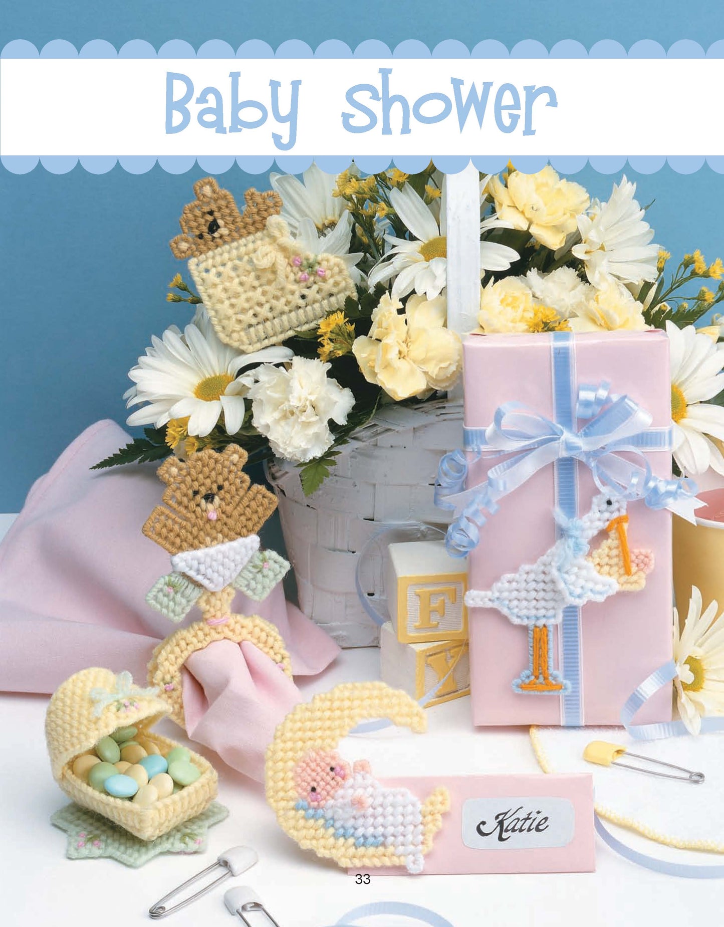 A pastel baby shower scene featuring crafts made with Friends for Baby in Plastic Canvas by Leisure Arts, plus a white basket of yellow and white flowers, shower favors, candies, baby blocks, a pink gift box with blue ribbon, and a Katie name card.