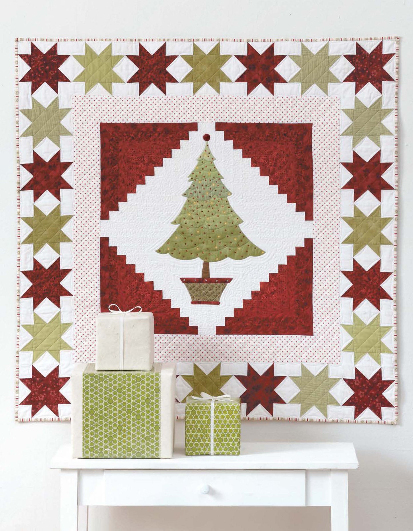 Add a festive touch to your decor with Leisure Arts’ Christmas Sparkle quilt, featuring a green tree with red and green star borders—plus seven whimsical embellishments that truly sparkle! Three wrapped gifts sit on a white table before the quilt.