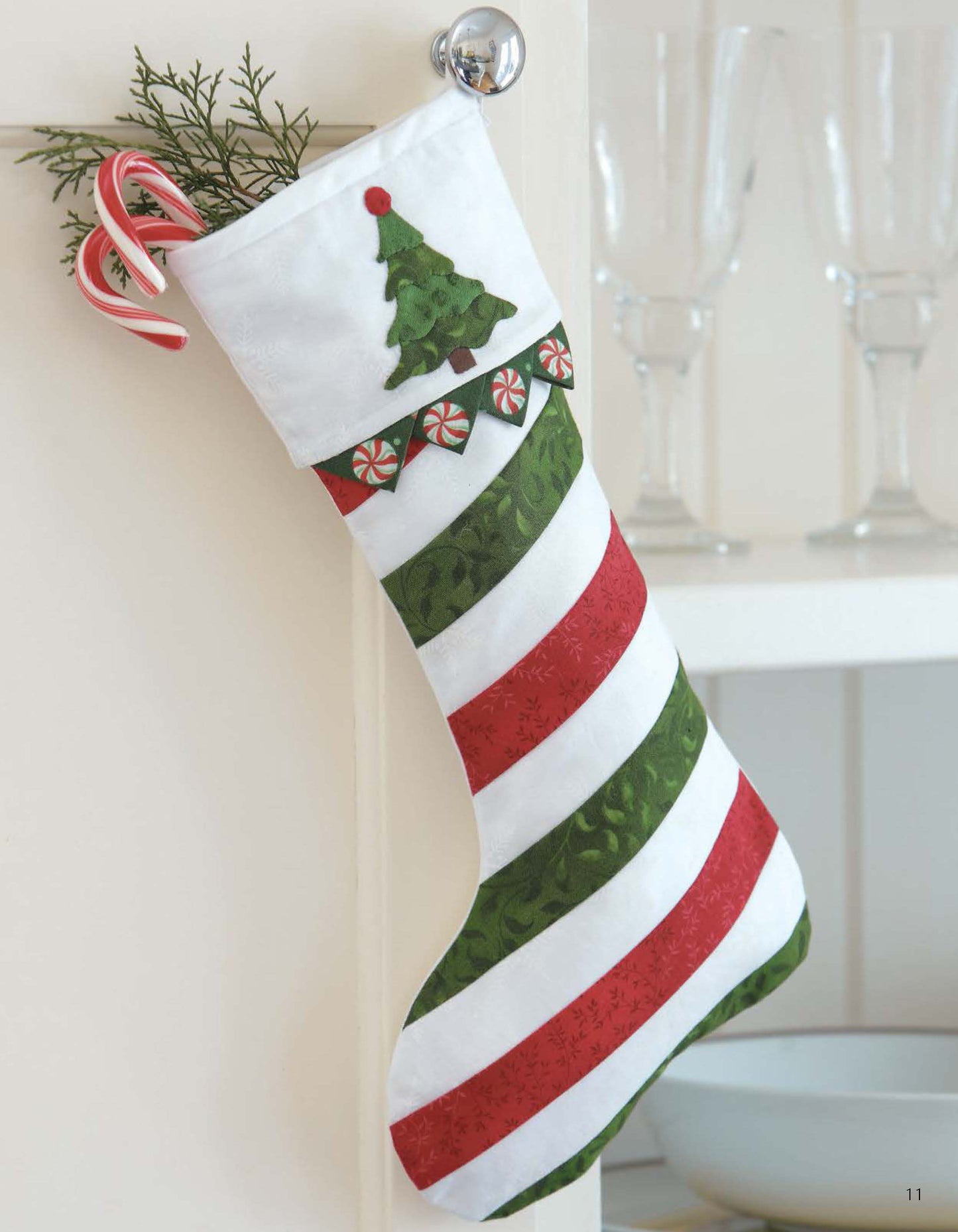 The Christmas Sparkle stocking by Leisure Arts, featuring 7 whimsical, sparkling embellishments, hangs on a knob. Filled with candy canes and greenery, it’s a festive decor accent. Glassware appears in the background.