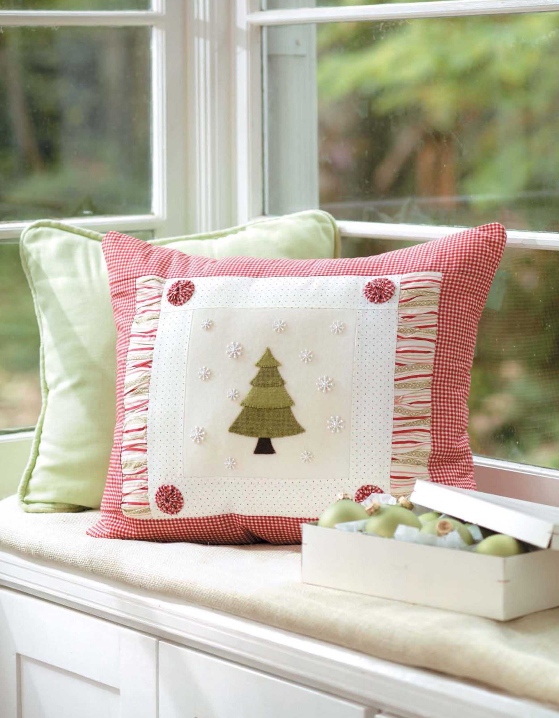 The Christmas Sparkle pillow by Leisure Arts features 7 whimsical, sparkling embellishments and red borders, placed on a window seat with light green cushions. A box of green apples adds charm to the festive décor.