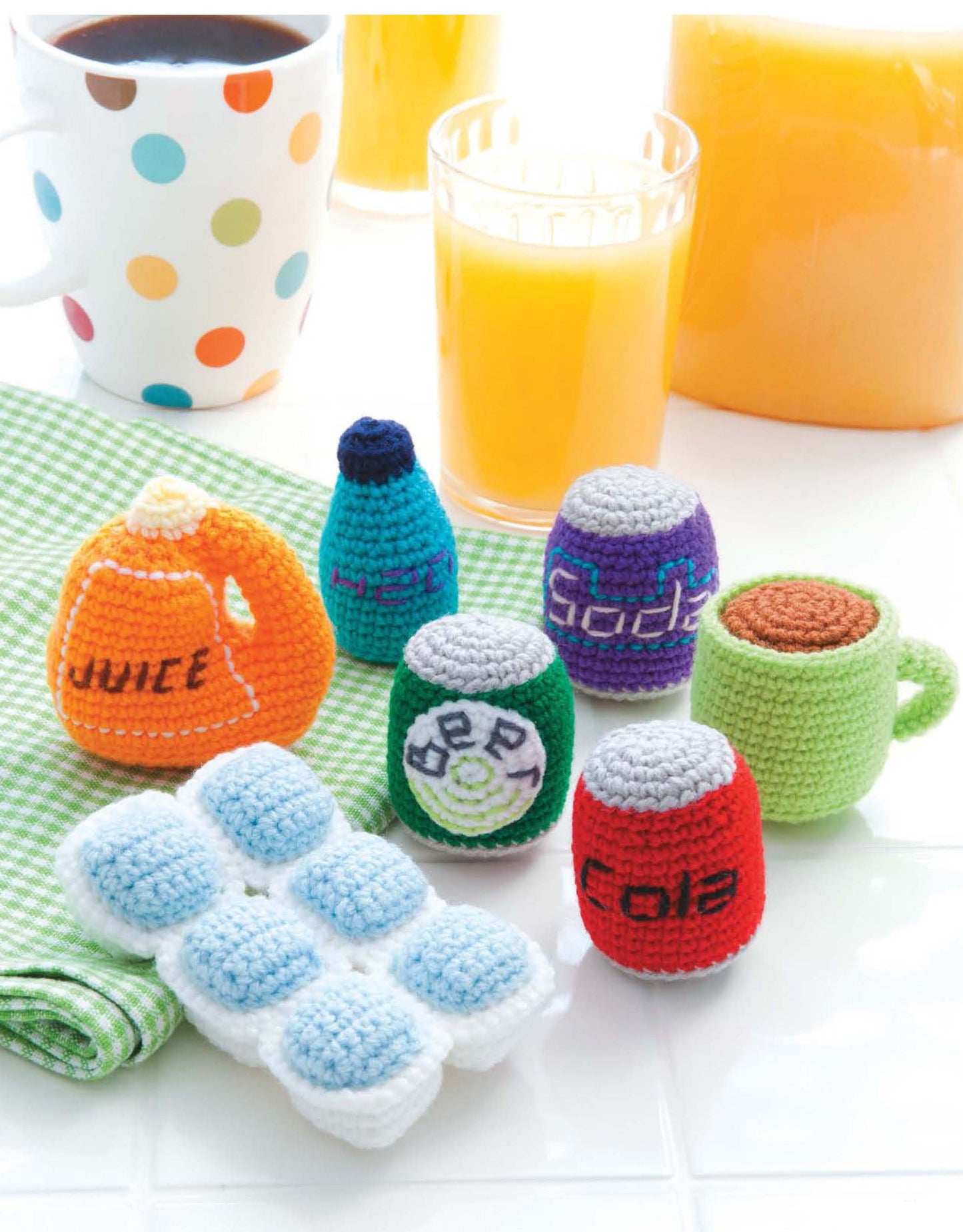 The Ice Box Crochet set by Leisure Arts creates a vibrant breakfast scene with crocheted faux foods—juice jug, cola, soda, mineral water, mug—and a white tray featuring crocheted ice cubes; real juice and coffee cups complete the look.